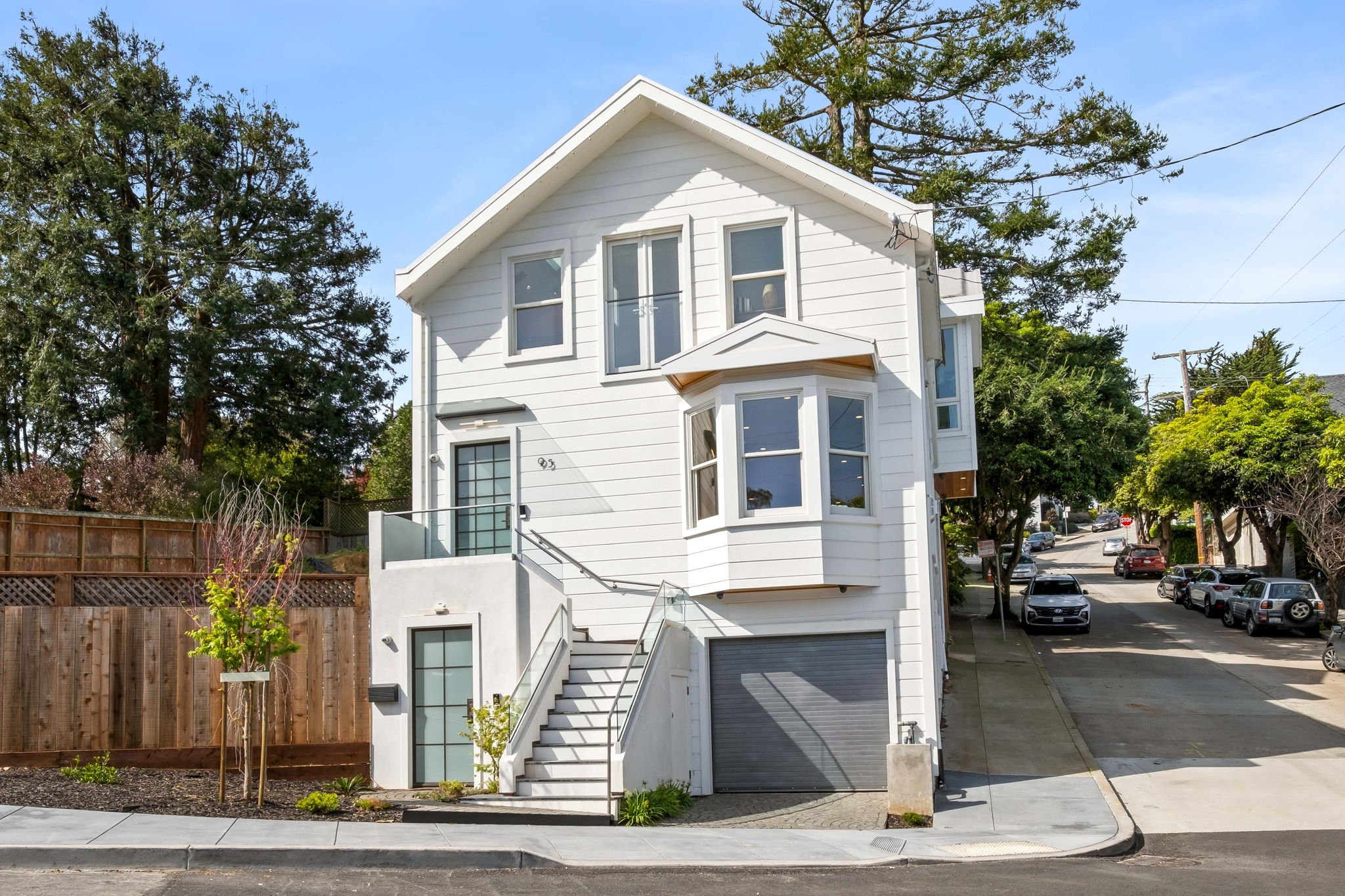 White three-story house with bay windows, staircase with glass railing, and a garage on a residential street with parked cars and trees.