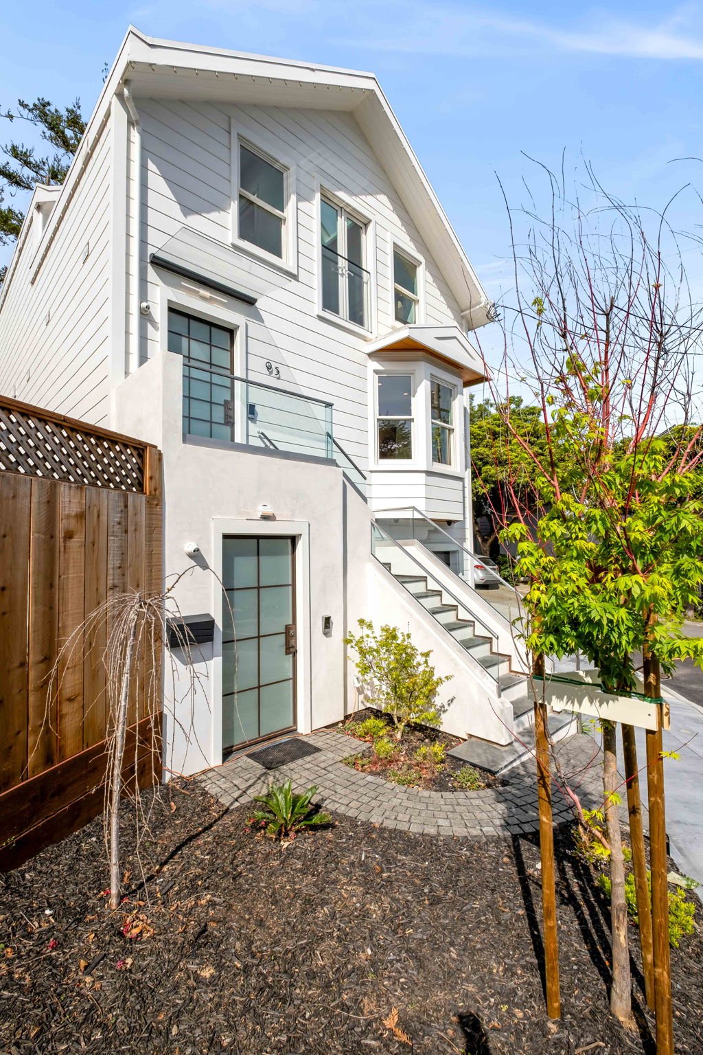 White multi-story house with outdoor staircase, balcony, and small landscaped yard with trees and plants.