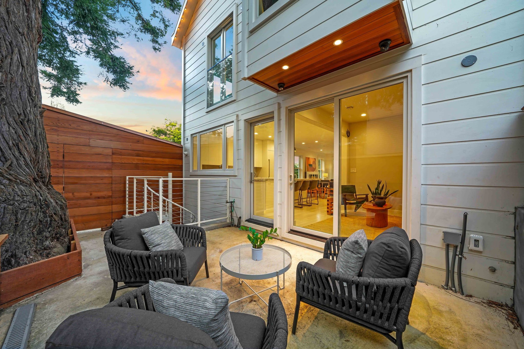 Residential backyard patio with black outdoor furniture, including two armchairs with gray pillows and a small round table with a potted plant, next to a large tree and a wooden privacy fence, with a view into the interior of a modern home through sl