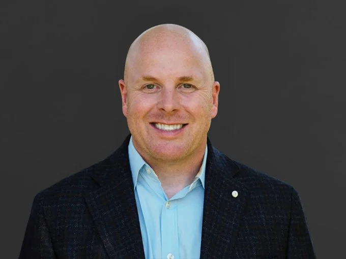A man with a bald head, smiling, wearing a dark blazer over a light blue dress shirt, posed against a plain gray background.