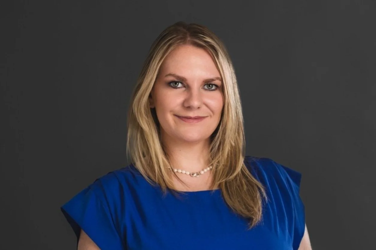 Portrait of a woman with blonde hair wearing a blue top and pearl necklace, standing against a dark gray background.