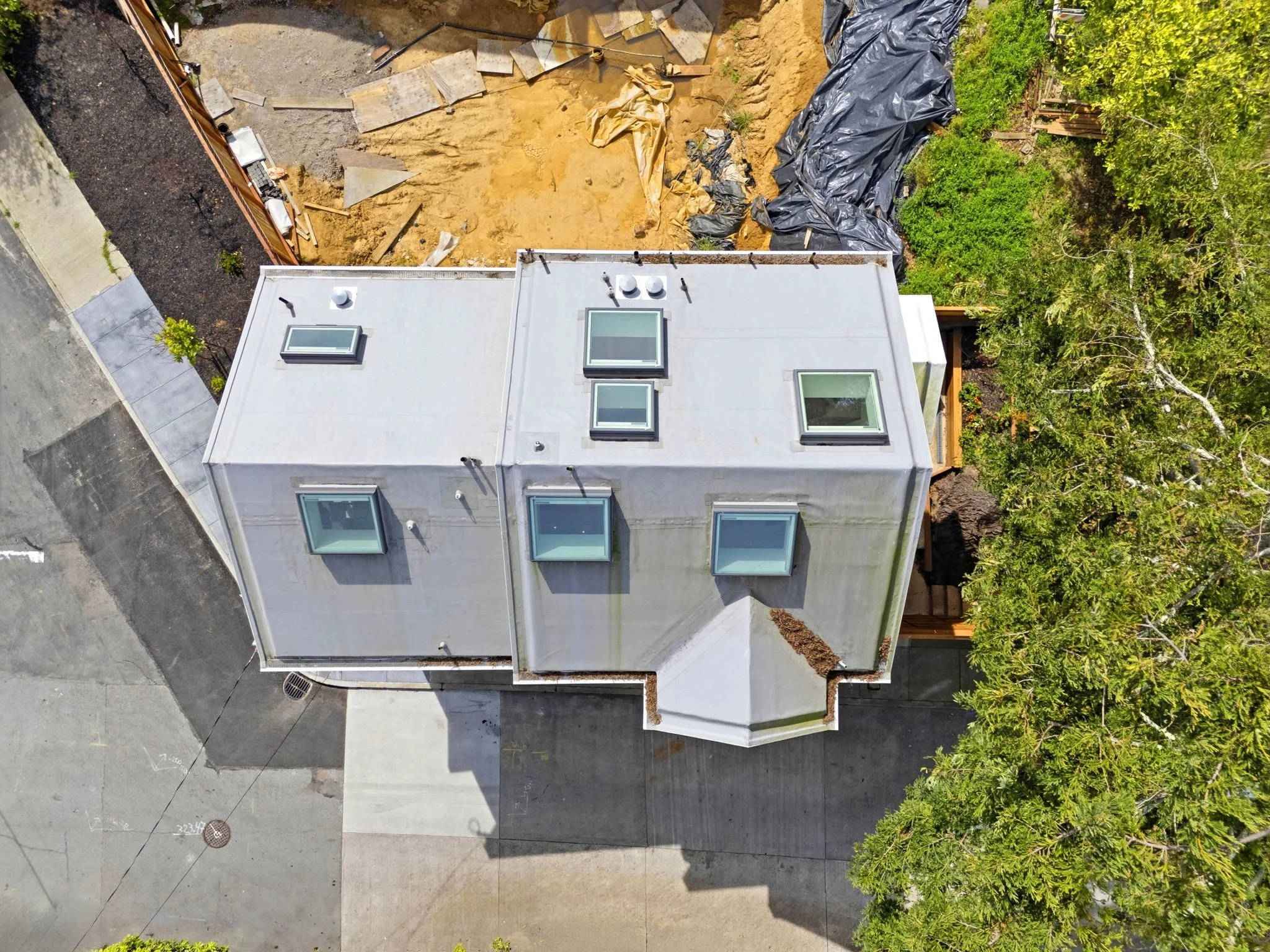 An aerial view of a house with a white roof, surrounded by trees, pavement, and a construction area with dirt and black plastic sheeting.