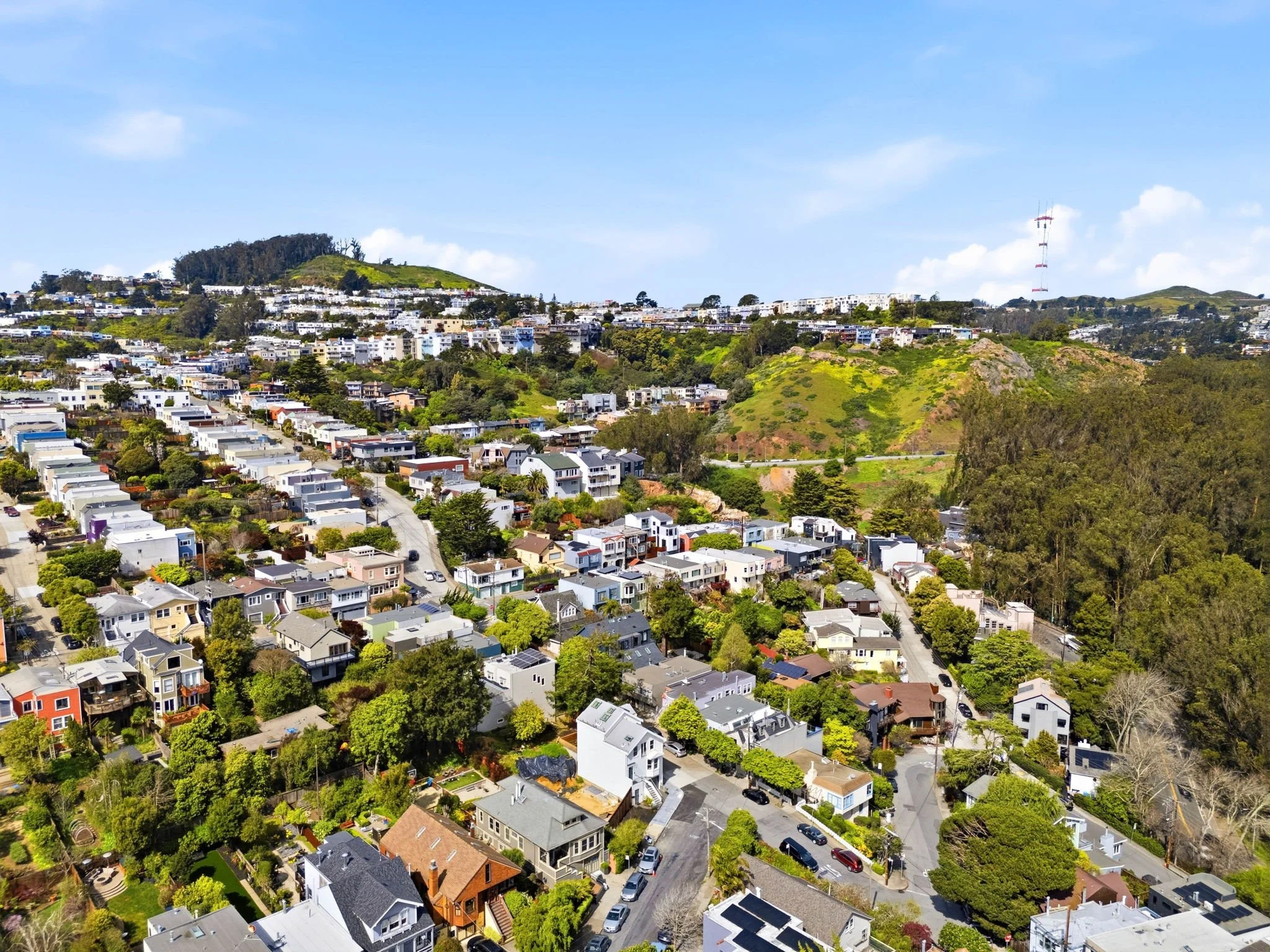 Aerial view of a hillside neighborhood with colorful houses, trees, and green hills in the background under a blue sky.