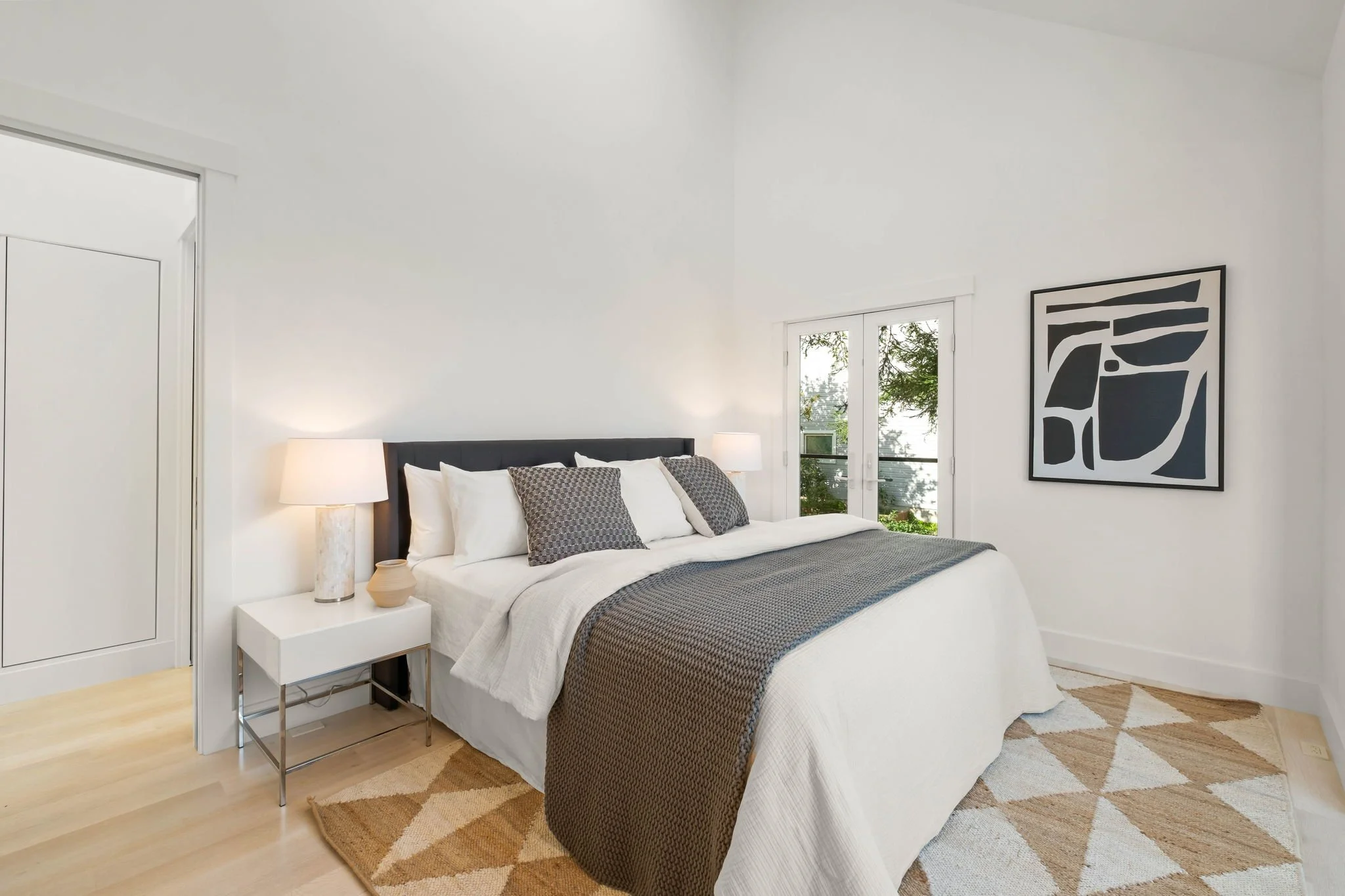 A neatly made bedroom with a white bed, gray blanket, decorative pillows, side table with lamps and vases, a geometric rug, wall art, and a glass door leading outside.