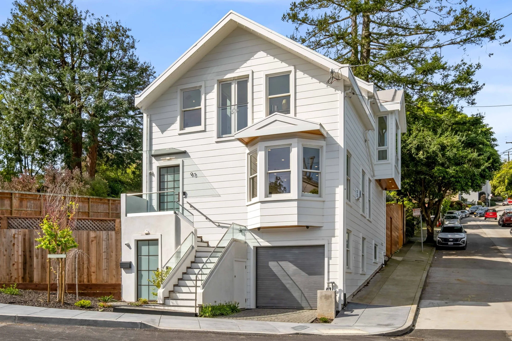 White multi-story house with a garage, staircase leading to the entrance, situated on a slightly elevated lot with trees and parked cars nearby.