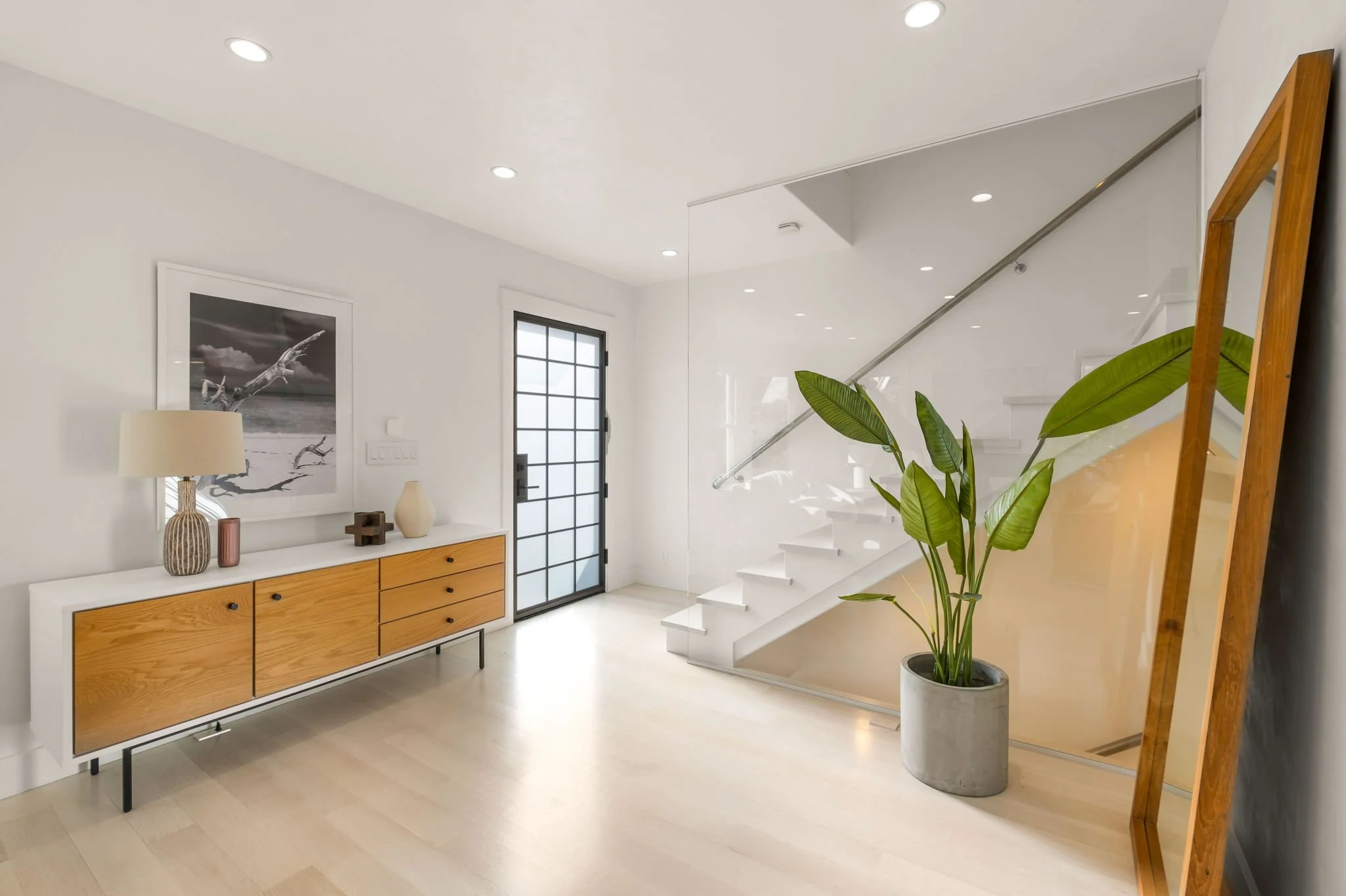 Modern minimalist interior with a white staircase, large potted plant, wooden console table with decorative items, black framed door, and black and white artwork on the wall