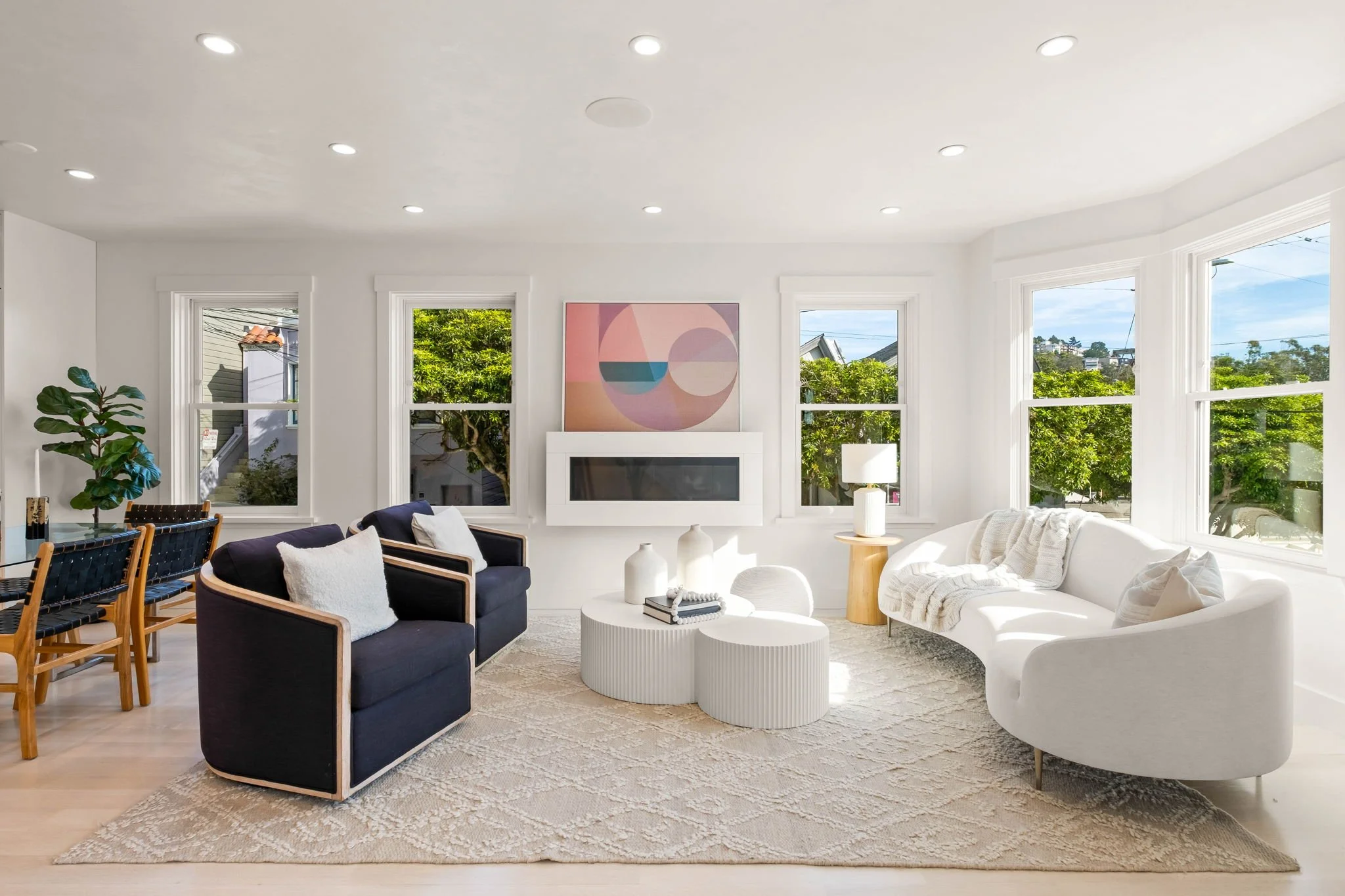 Bright living room with white walls, multiple windows showing greenery outside, a white sofa, two navy armchairs with white pillows, a white textured rug, and white and wooden side tables. There is a modern fireplace and a colorful abstract painting on the wall above it.