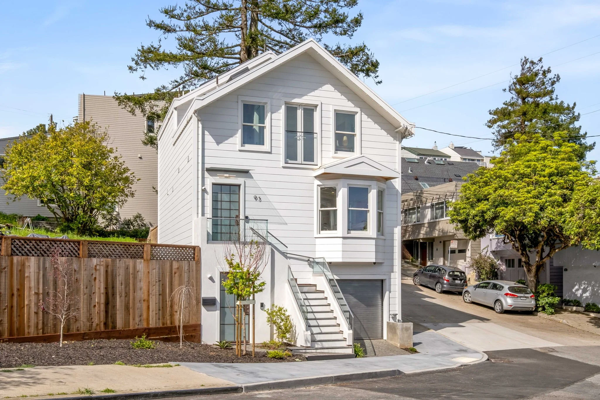 A white multi-story house with stairs leading to the front door, located on a sloped street, with trees and parked cars in the background.