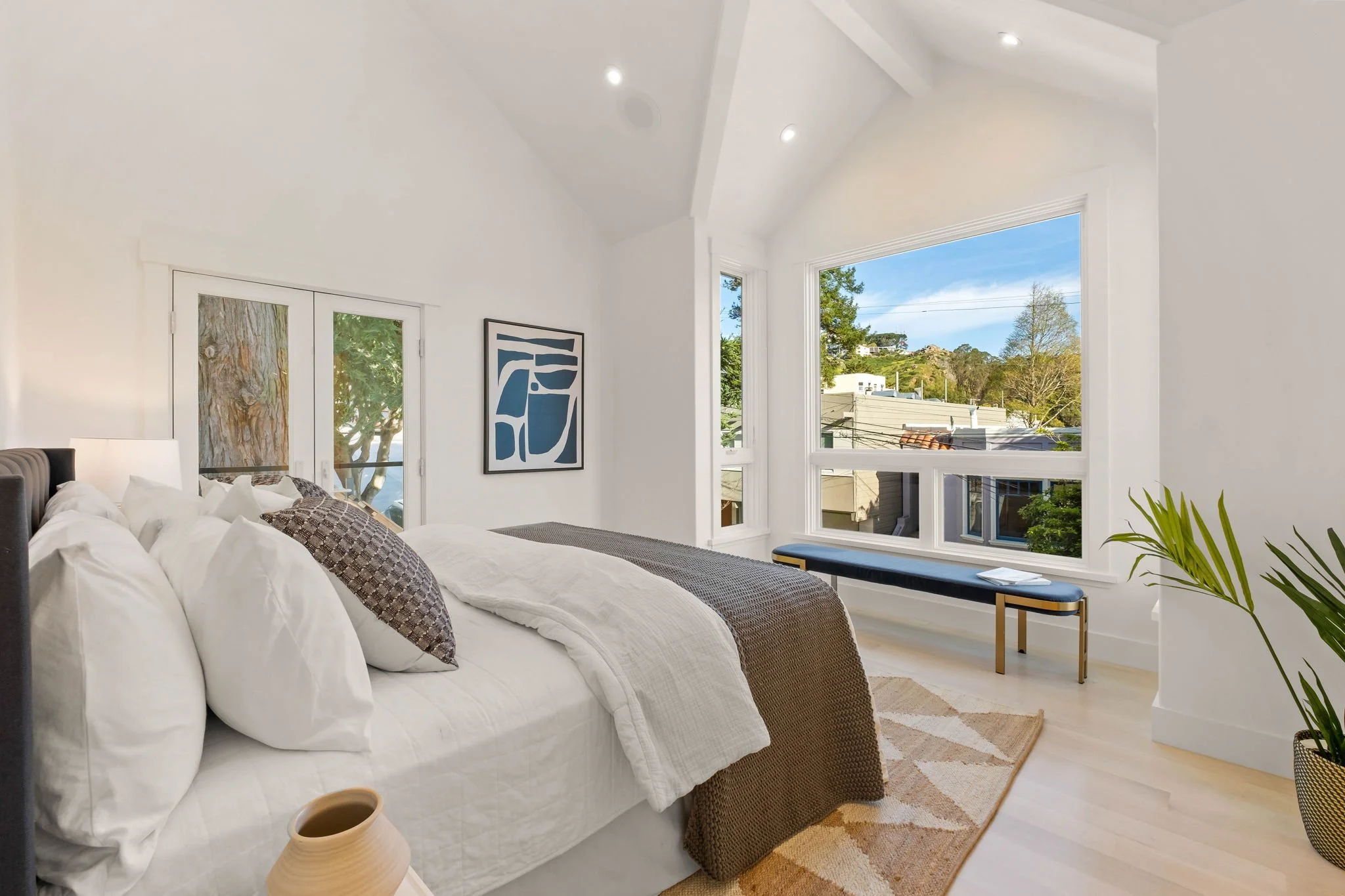 Bright and airy bedroom with white walls and large windows showing a view of trees and buildings outside. The bed has white linens and patterned pillows. A bench is placed under the windows, and there's a potted plant in the foreground.