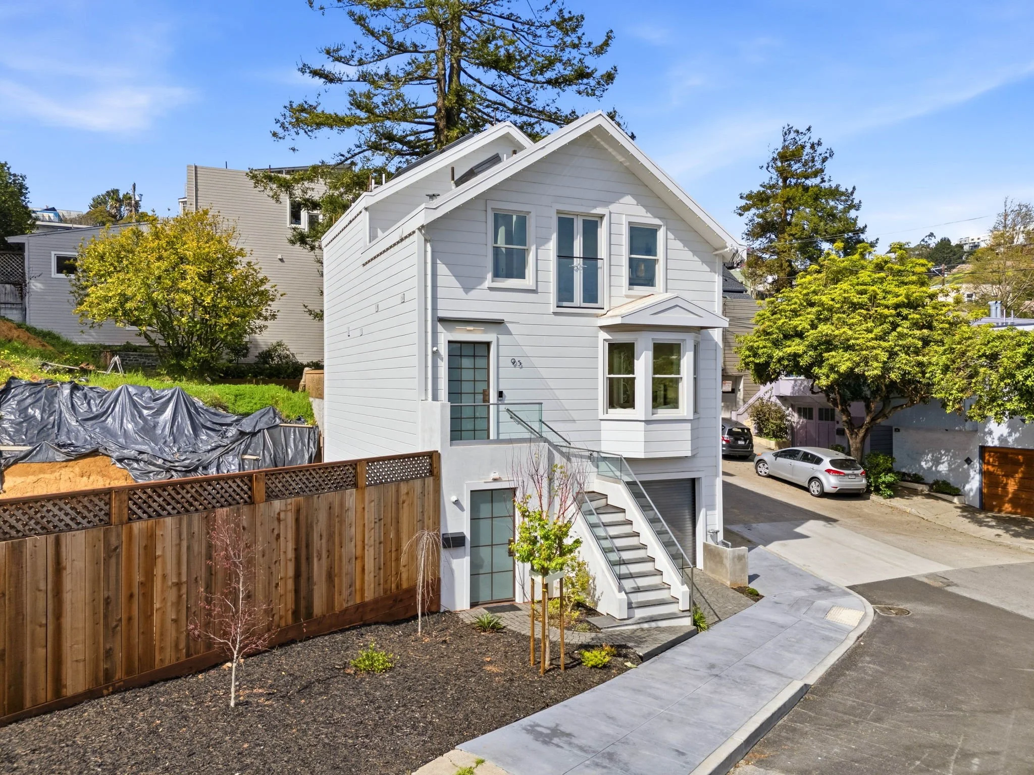 A modern white house with three floors, located on a curved street, with a front staircase, greenery, and parked cars nearby.
