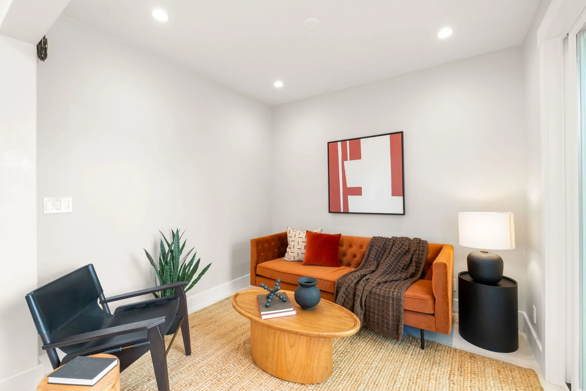A modern living room with white walls, a tan rug, an orange tufted sofa with throw pillows and a blanket, a black side table with a lamp, a black armchair, a wooden coffee table with decorative objects, an abstract art piece on the wall, and a potted