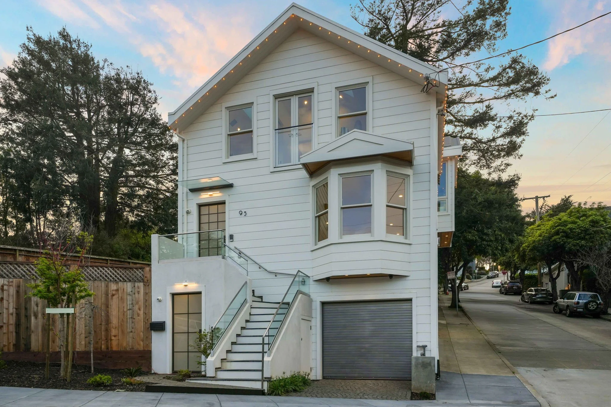 A modern three-story white house with large windows, exterior lighting, and a garage on a street at sunset.