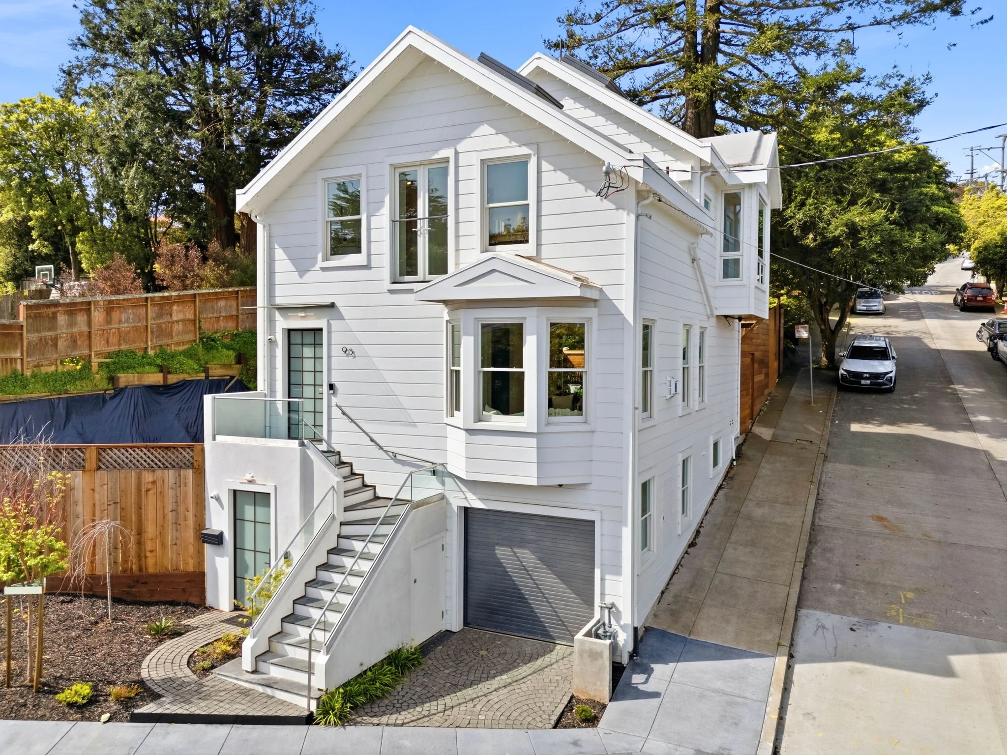 White multi-story house with stairs leading to front door, situated on a slight hill, with a garage at the bottom, on a city street with three cars parked.