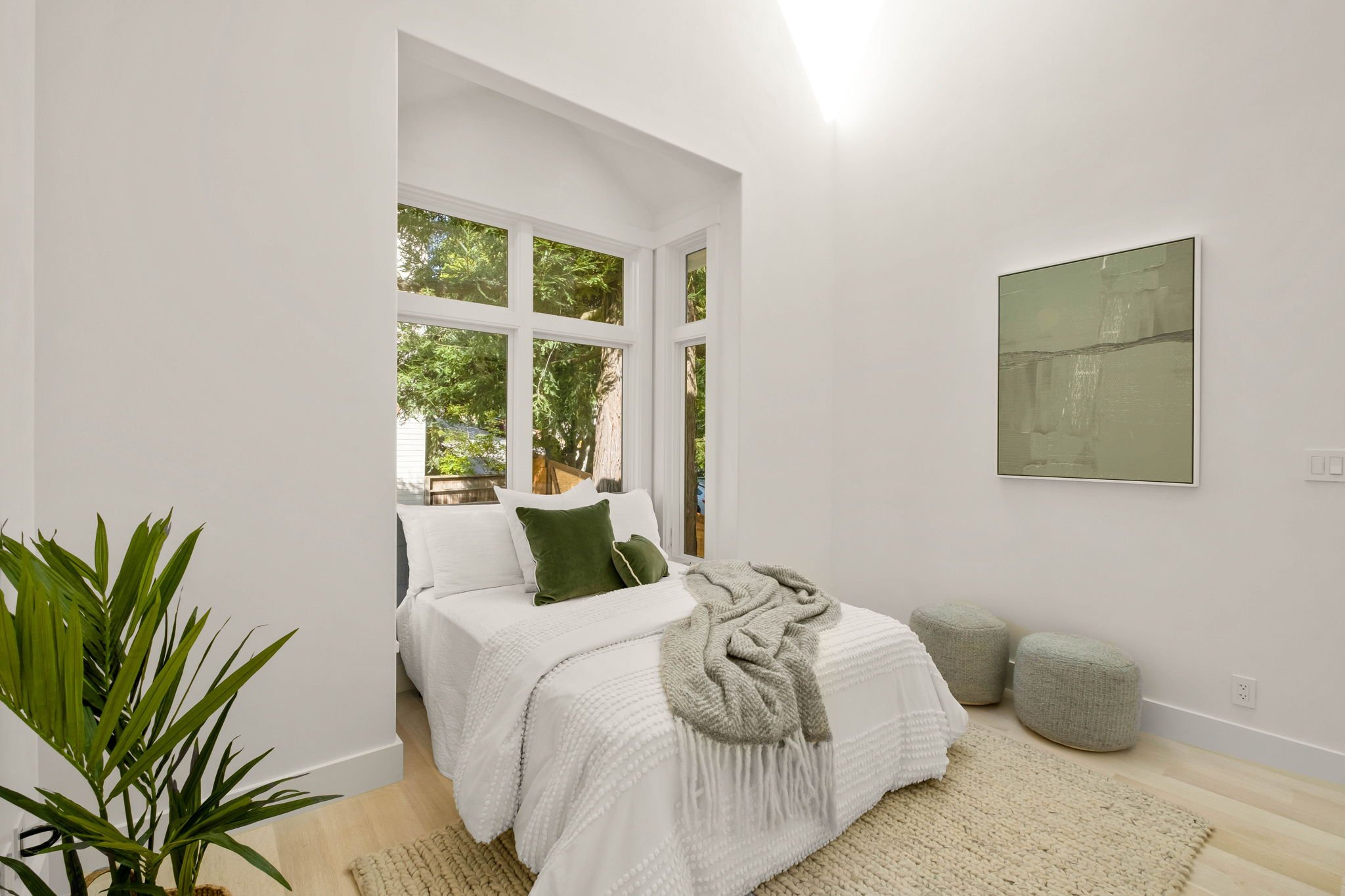 A bright, minimalist bedroom with a large window, white walls, and white bedding accented with green pillows. Soft gray poufs and a textured beige rug complete the cozy and airy space.