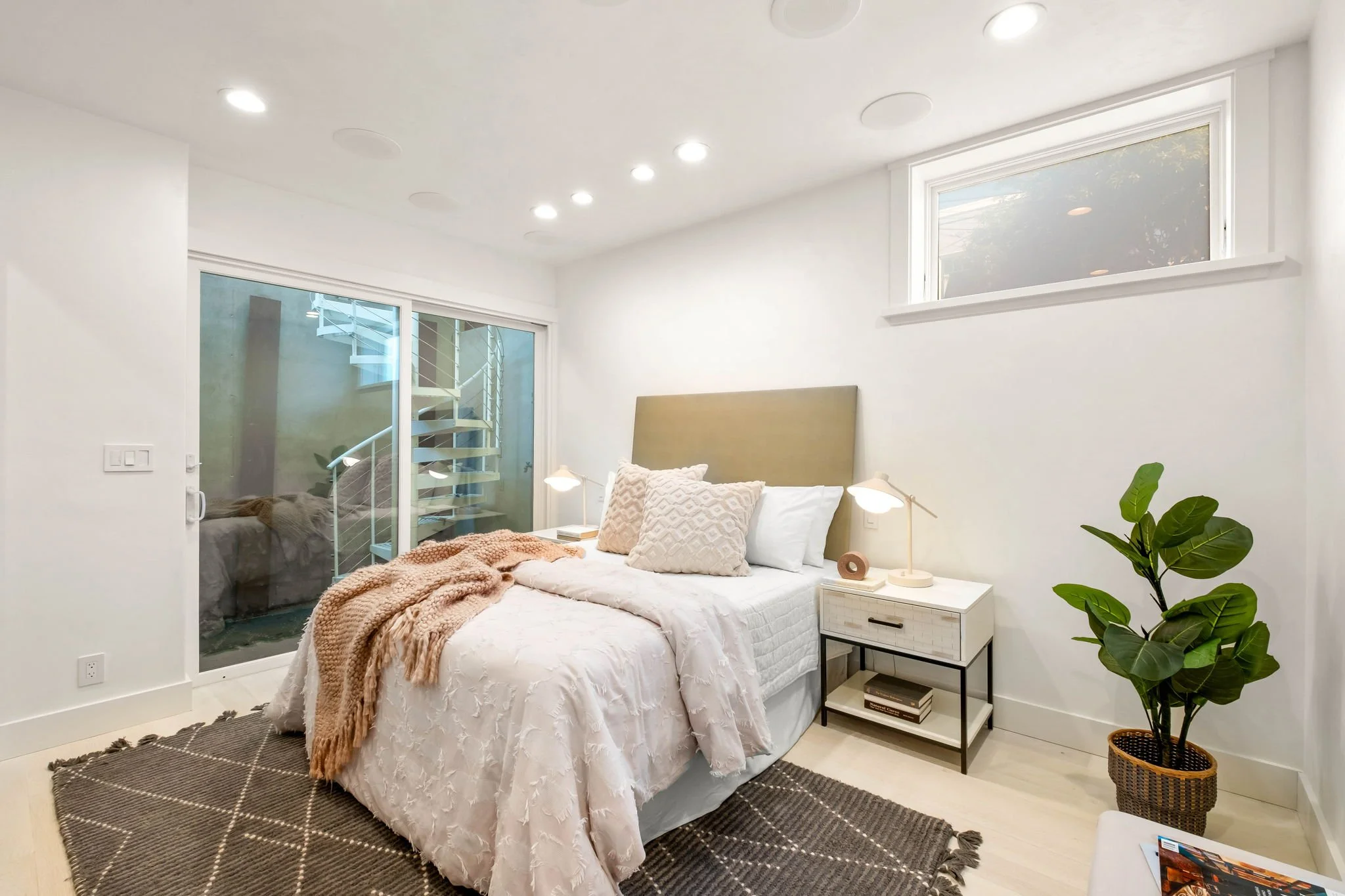 A modern bedroom with a neatly made bed, beige headboard, white bedding, and decorative pillows. There are two bedside lamps on white nightstands, a potted plant in the corner, a wooden floor, a brown rug, and a sliding glass door leading to a stairc