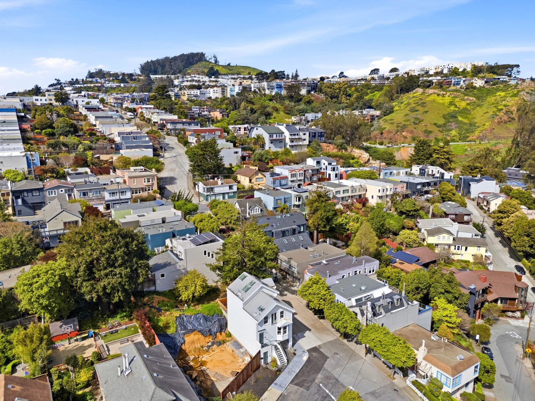 Aerial view of a hillside residential neighborhood with colorful houses, trees, and winding streets under a partly cloudy sky.
