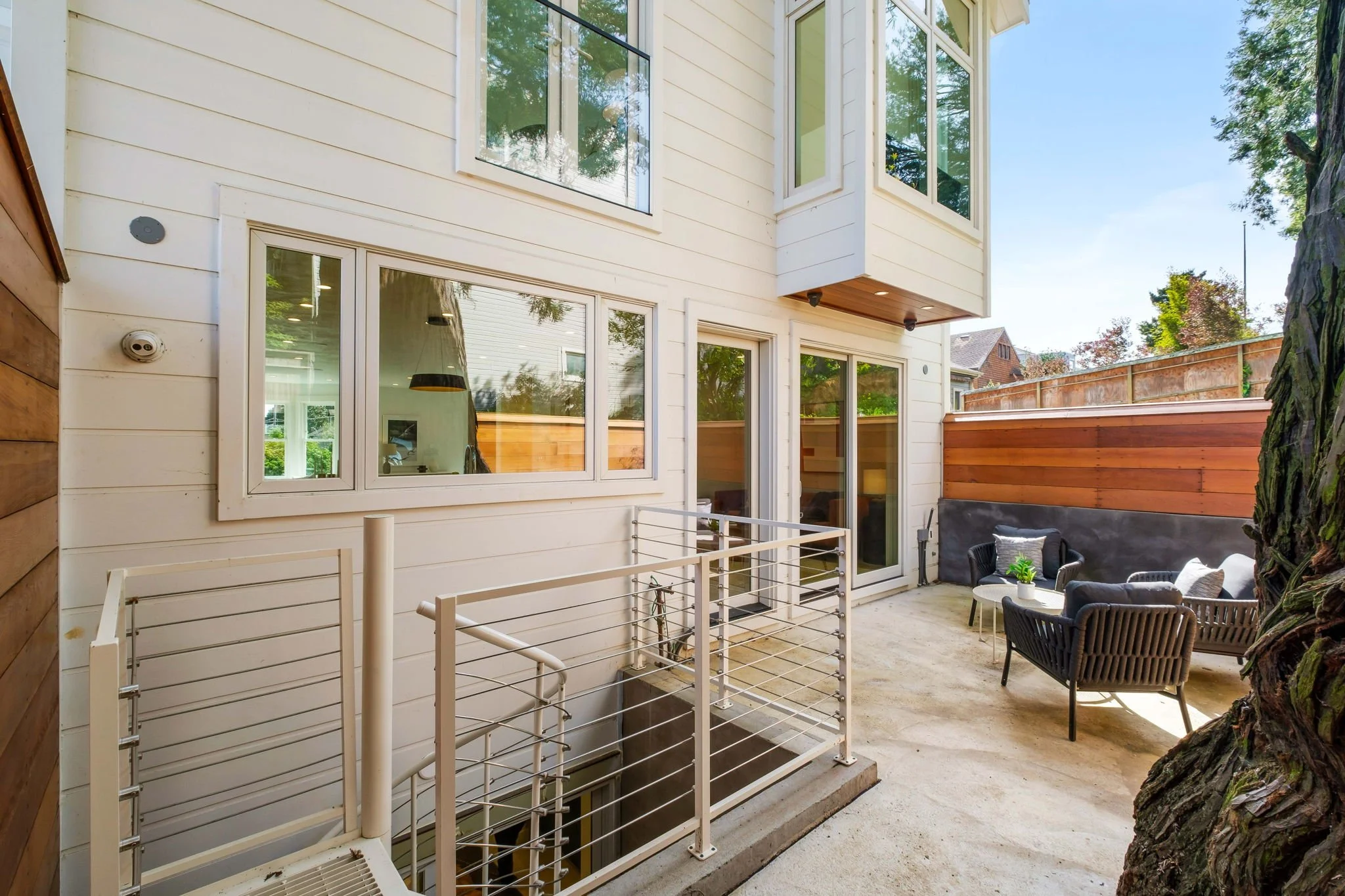 Outdoor patio with a seating area featuring black chairs and a small table, surrounded by a wooden fence and large windows on a white house, with trees visible in the background.