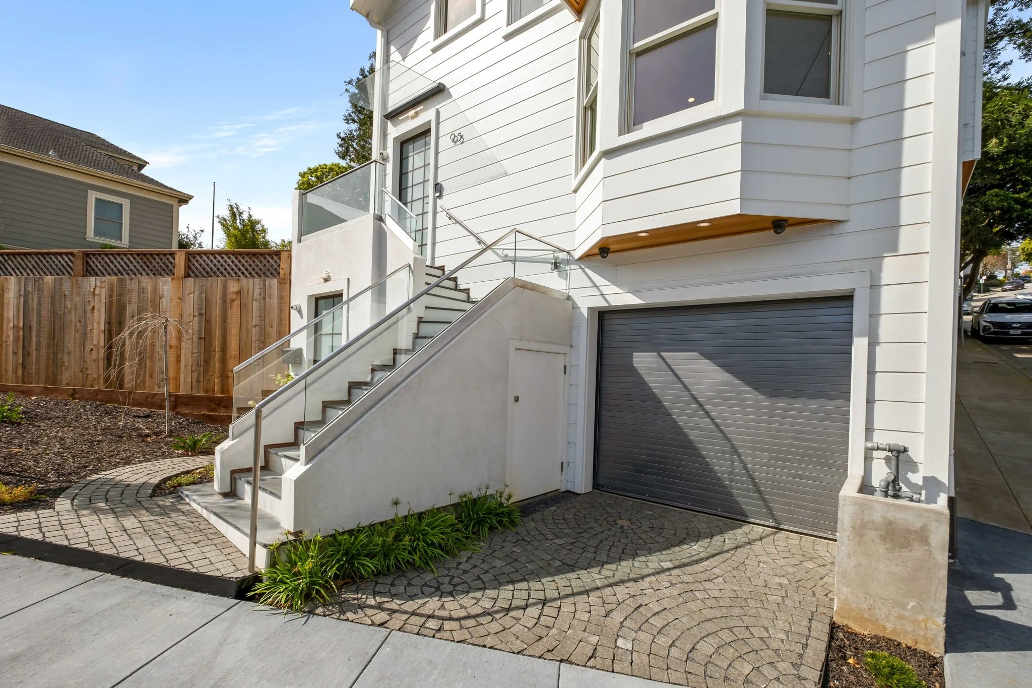 Exterior view of a modern white house with stairs leading to an upper level, a garage door beneath, and a small landscaped area with green plants in the front.