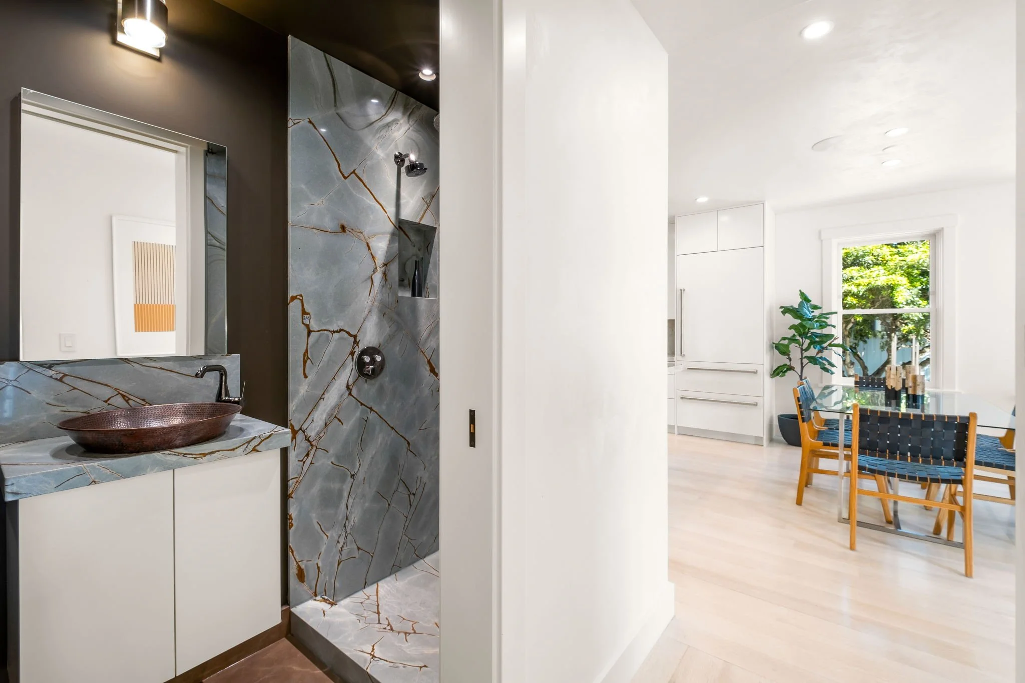 Interior view of a modern home with visible bathroom vanity area featuring marble surfaces and a copper sink, and a dining area with a table, chairs, a large window, and a potted plant.
