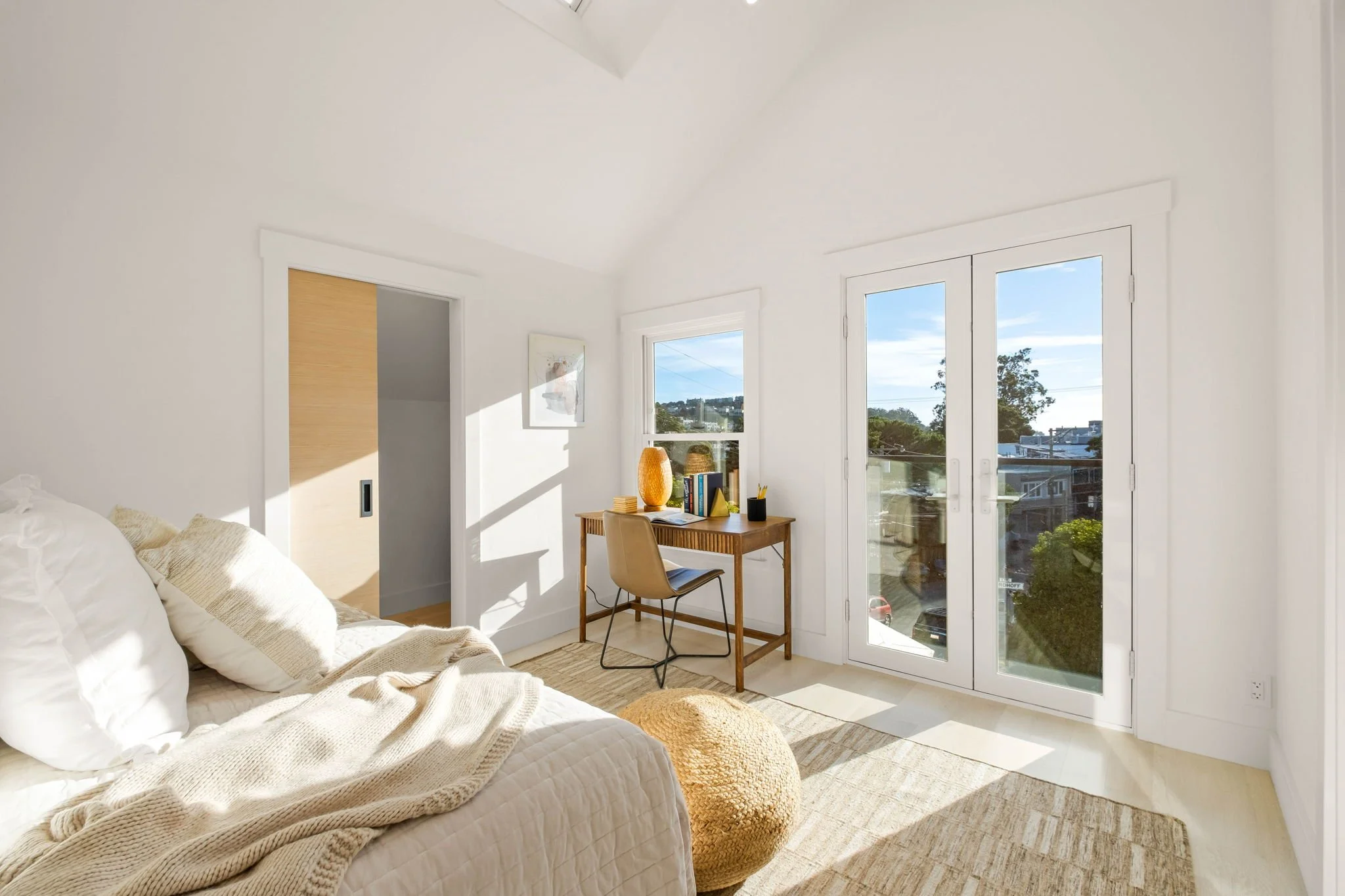 Bright bedroom with white walls, a bed with white bedding and pillows, a round beige pouf, a wooden desk with books, a decorative vase, and a chair. Large windows and glass door show a sunny outdoor view with trees and buildings.