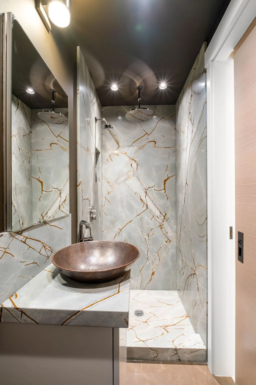 A modern bathroom with marble walls and a marble sink, featuring two rain showerheads on the ceiling and a mirror above the sink.