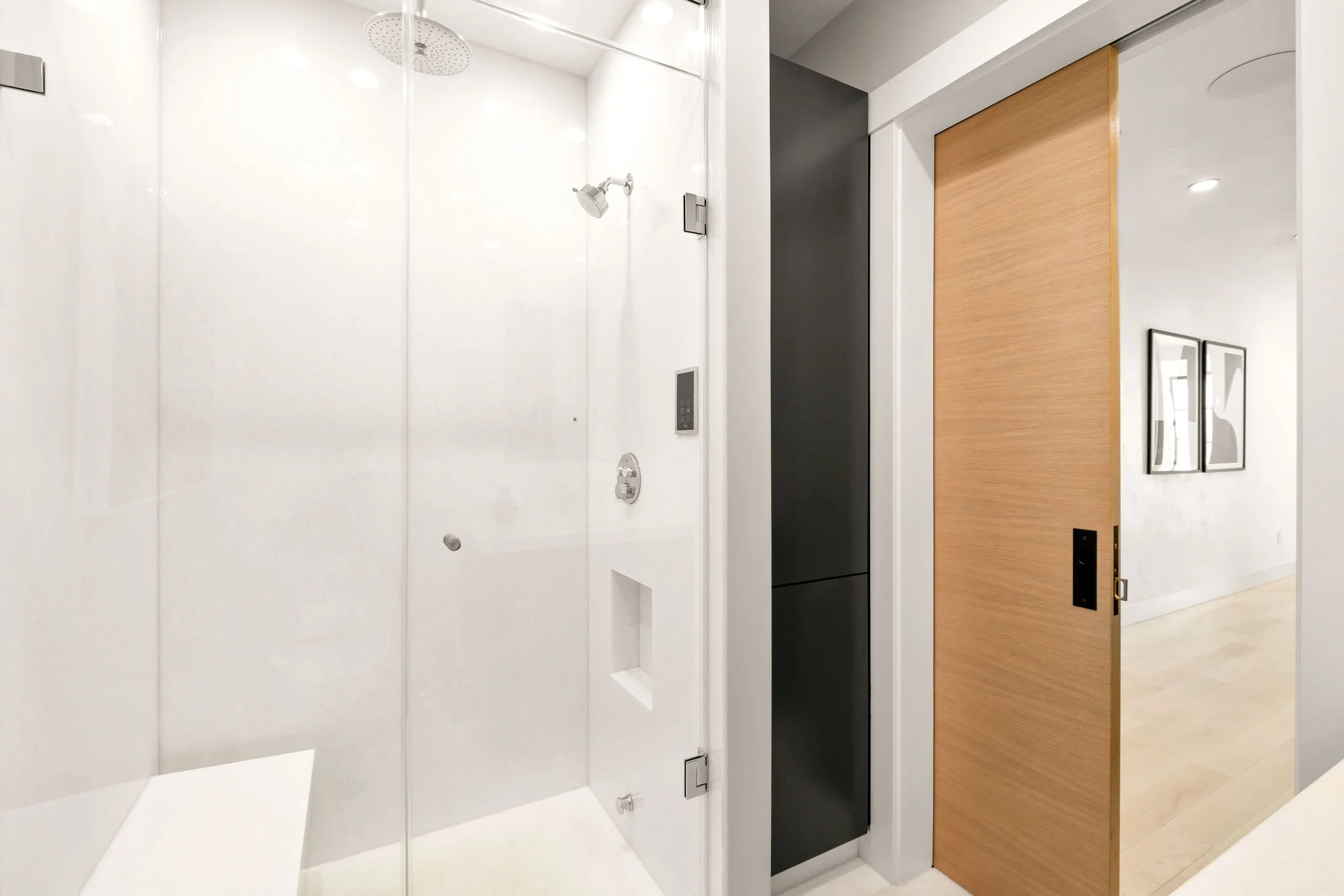 Modern shower area with a glass door, rainfall showerhead, and handheld shower, adjacent to a sliding wooden door revealing a hallway with framed artwork.