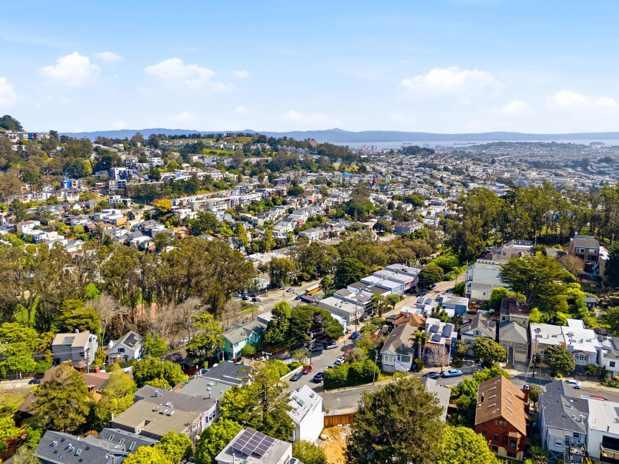 Aerial view of a densely populated hillside neighborhood with residential buildings, trees, and streets, overlooking a bay and distant hills under a blue sky with scattered clouds.