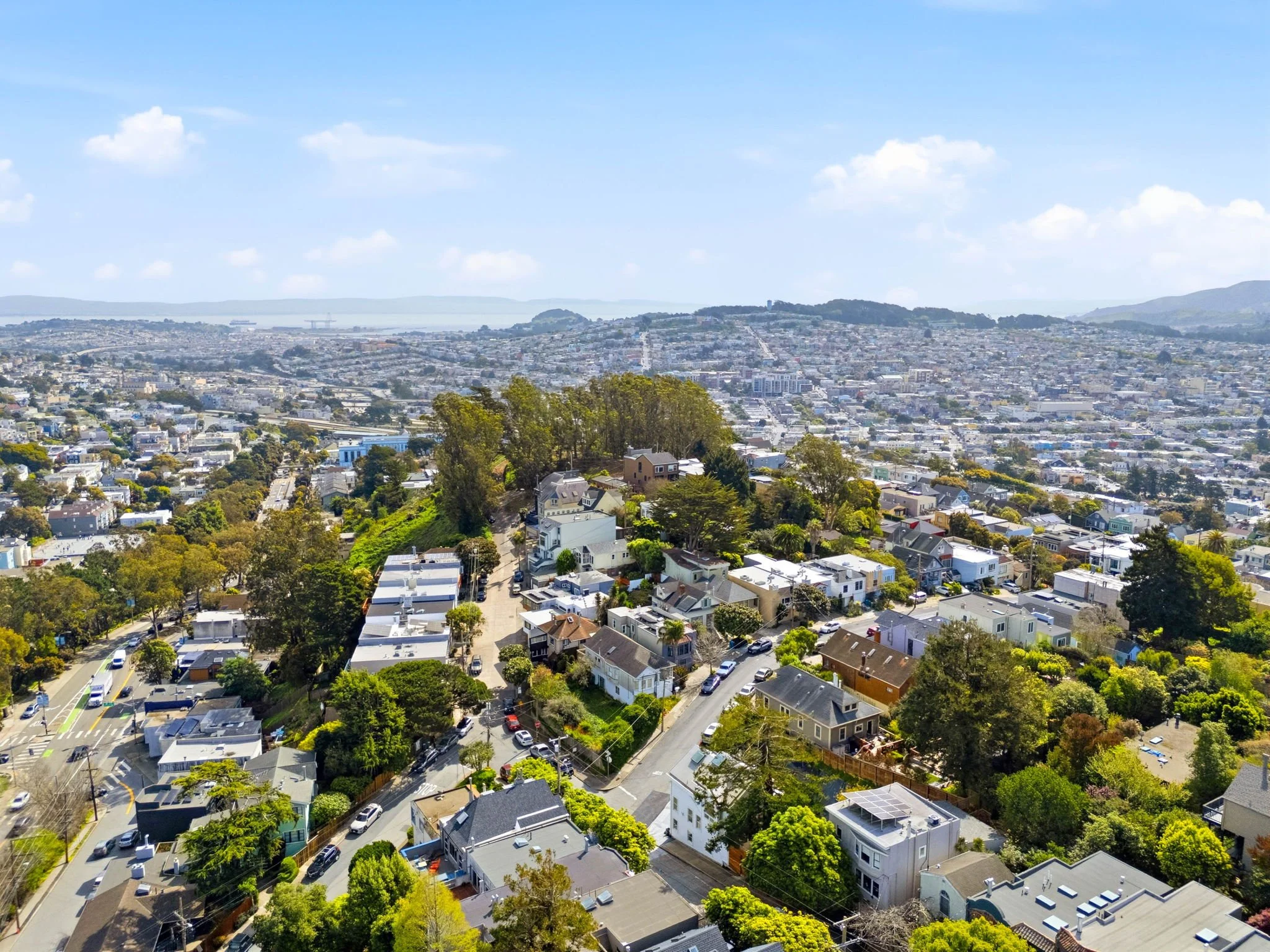 Aerial view of a residential neighborhood with houses, trees, and streets, with a cityscape and hills in the background under a partly cloudy sky.