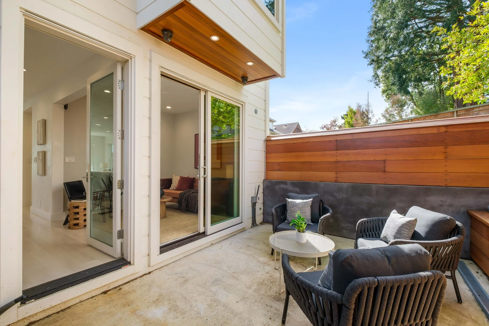 Outdoor patio with black wicker chairs and a small white coffee table with a potted plant, enclosed by a wooden privacy wall, adjacent to a house with large sliding glass doors leading inside.