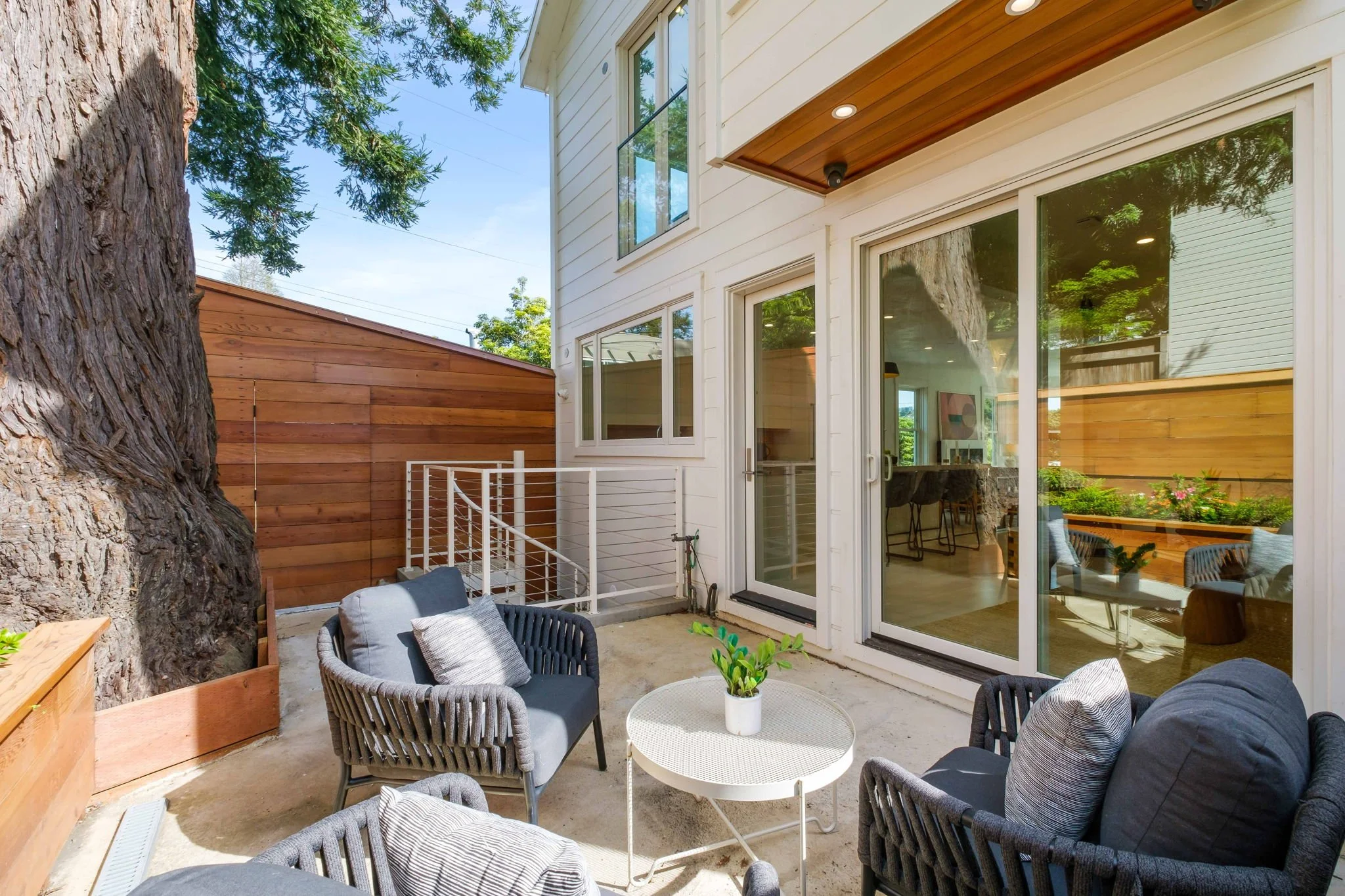 Outdoor patio with black chairs and a white round table with a potted plant, adjacent to a white house with large windows and sliding glass door, surrounded by tall trees and wooden fence.