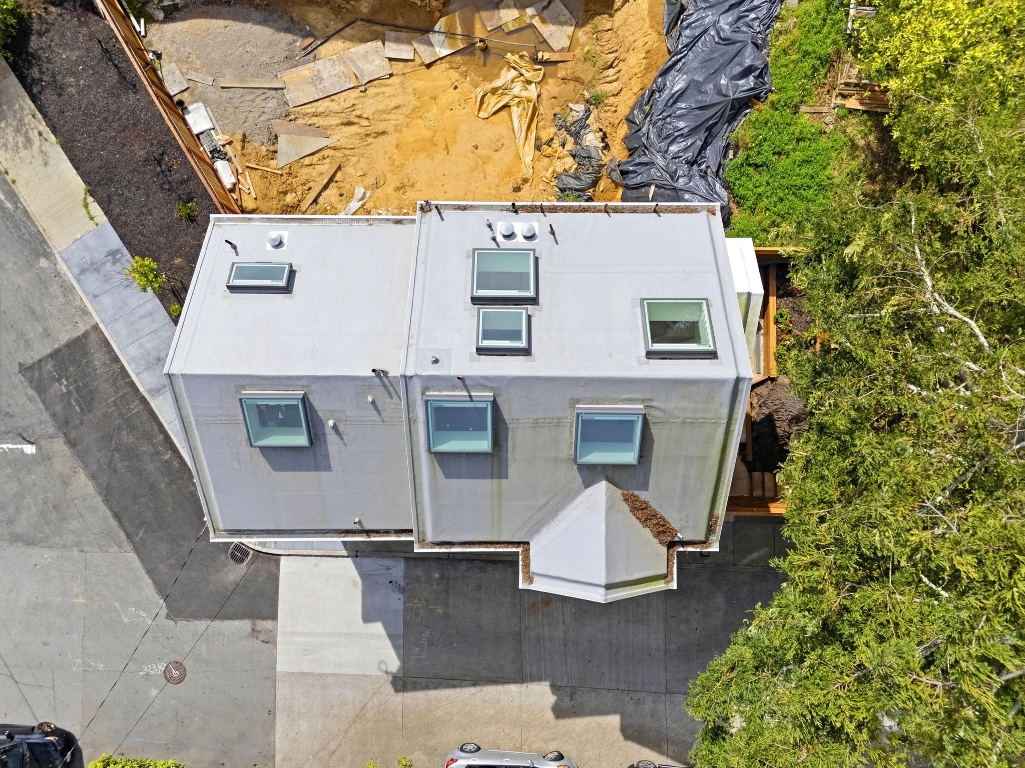 An aerial view of a modern house with a flat roof and multiple skylights, surrounded by a paved driveway, trees, and a construction site with dirt and black plastic covering.