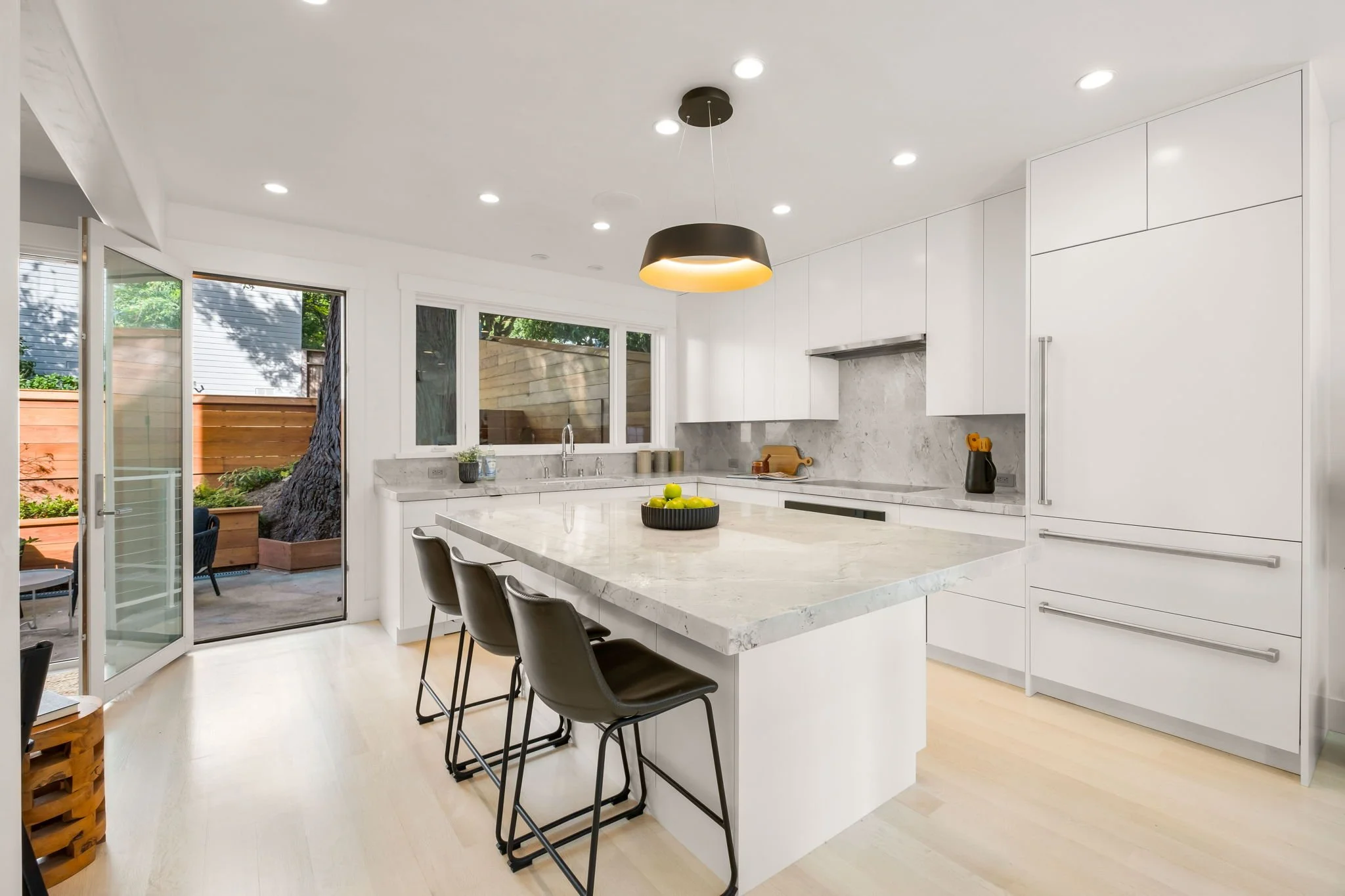 Modern white kitchen with marble countertops, black bar stools, and large windows opening to an outdoor patio with trees and wooden fencing.