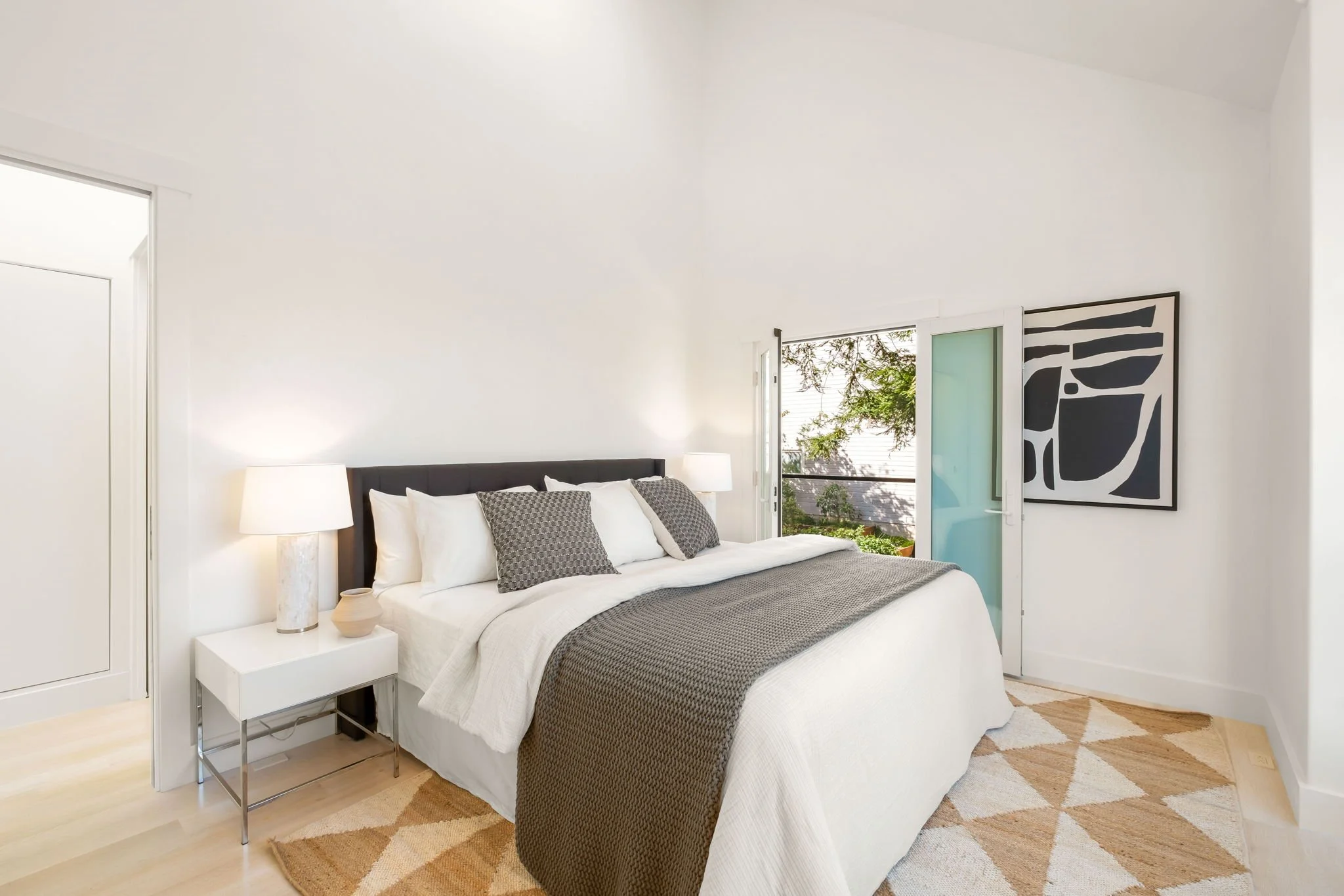 Bright bedroom with a white bed, dark headboard, gray pillows, and blanket, side table with lamp and decorative object, sliding glass door with a balcony, artwork on wall, and a geometric patterned rug.