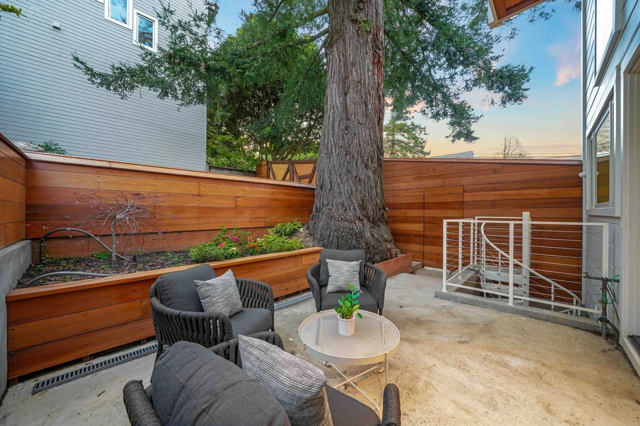 Backyard patio with a large tree, wooden fencing, outdoor seating with pillows, a small round table with a plant, and a staircase leading down.