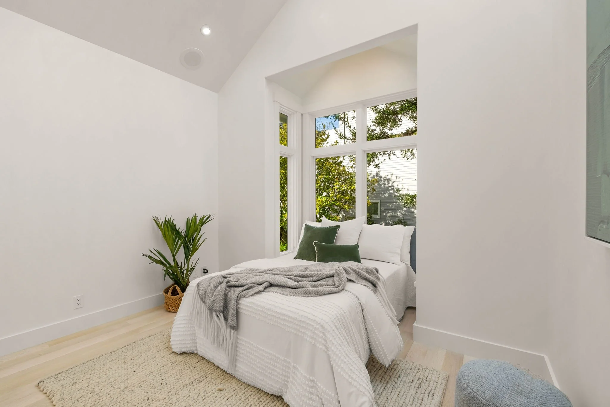 A cozy bedroom with white bedding, green accent pillows, a gray blanket, and a large window with greenery outside.