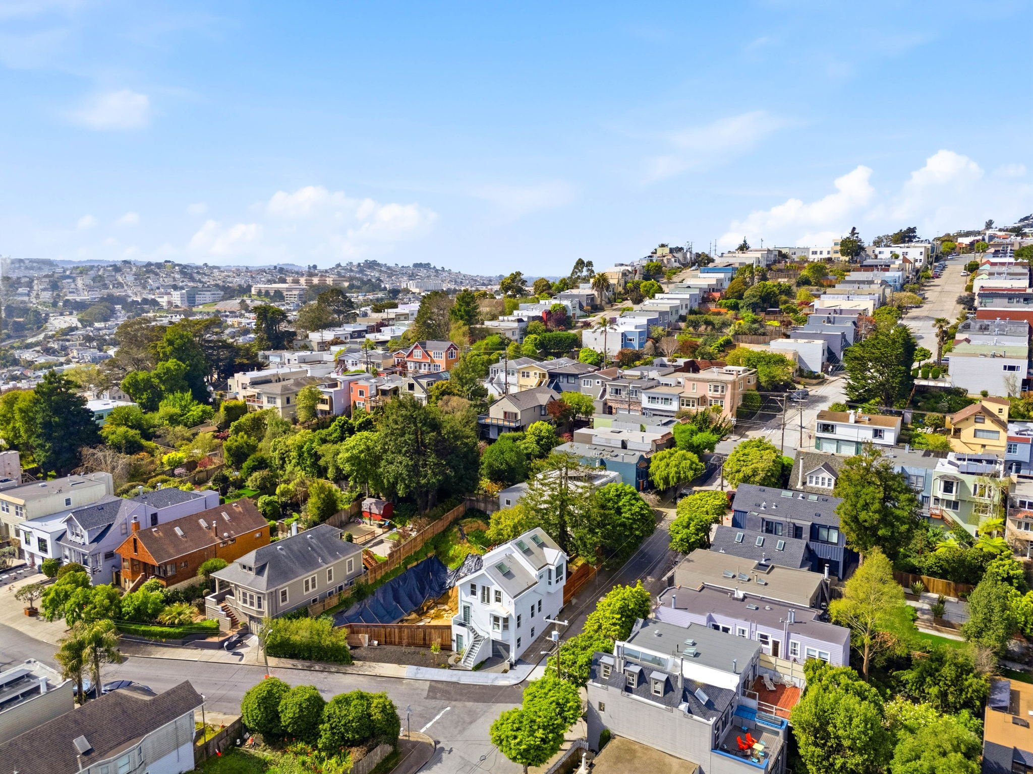 Aerial view of a hilly residential neighborhood with colorful houses, lush green trees, and streets winding uphill on a sunny day with a partly cloudy sky.