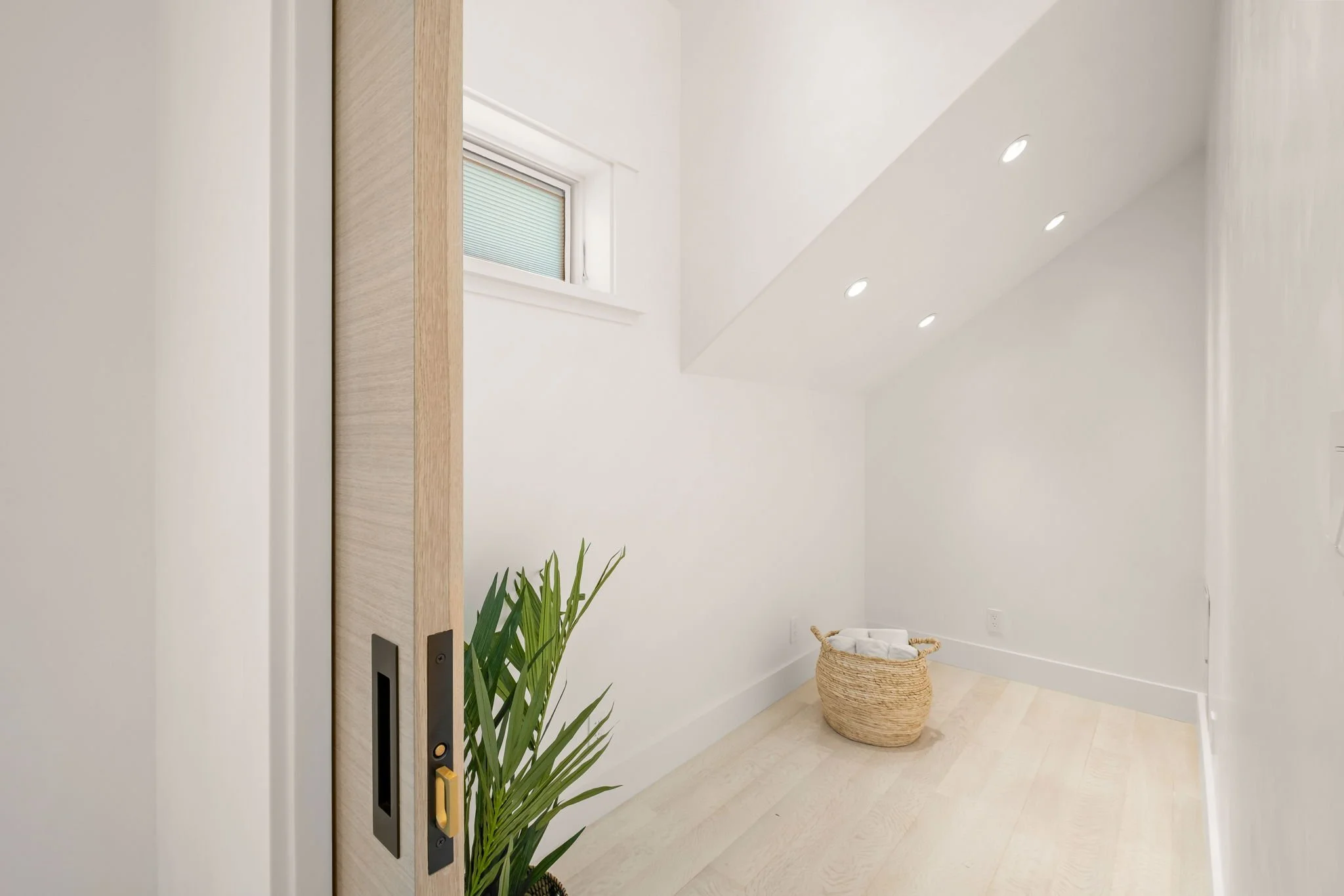 Minimalist room with white walls, a small window, recessed ceiling lights, a woven basket with folded towels, and a green plant near a sliding wooden door.