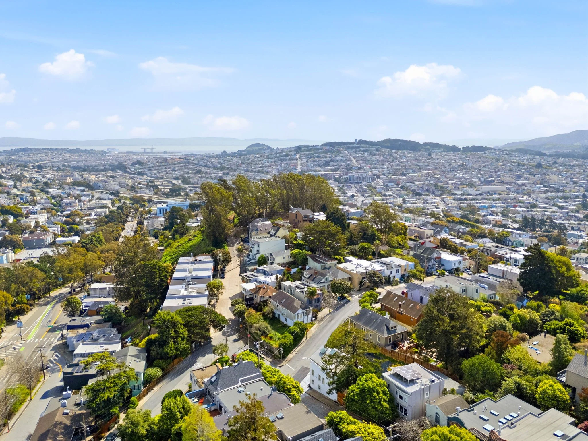 Aerial view of a hilly residential area with houses, trees, and streets, with a cityscape and distant water in the background under a partly cloudy sky.
