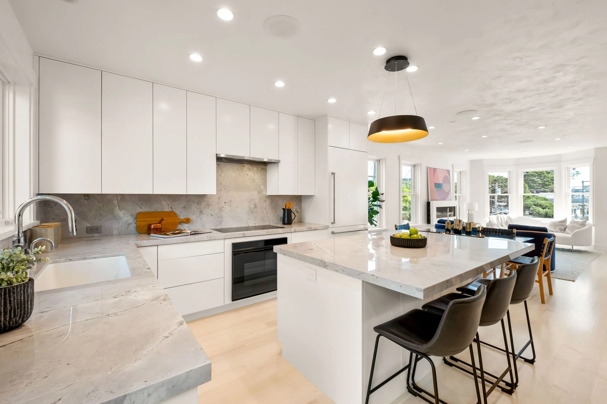 Modern white kitchen with marble countertops, an island with seating, and a view into a bright living room with large windows.