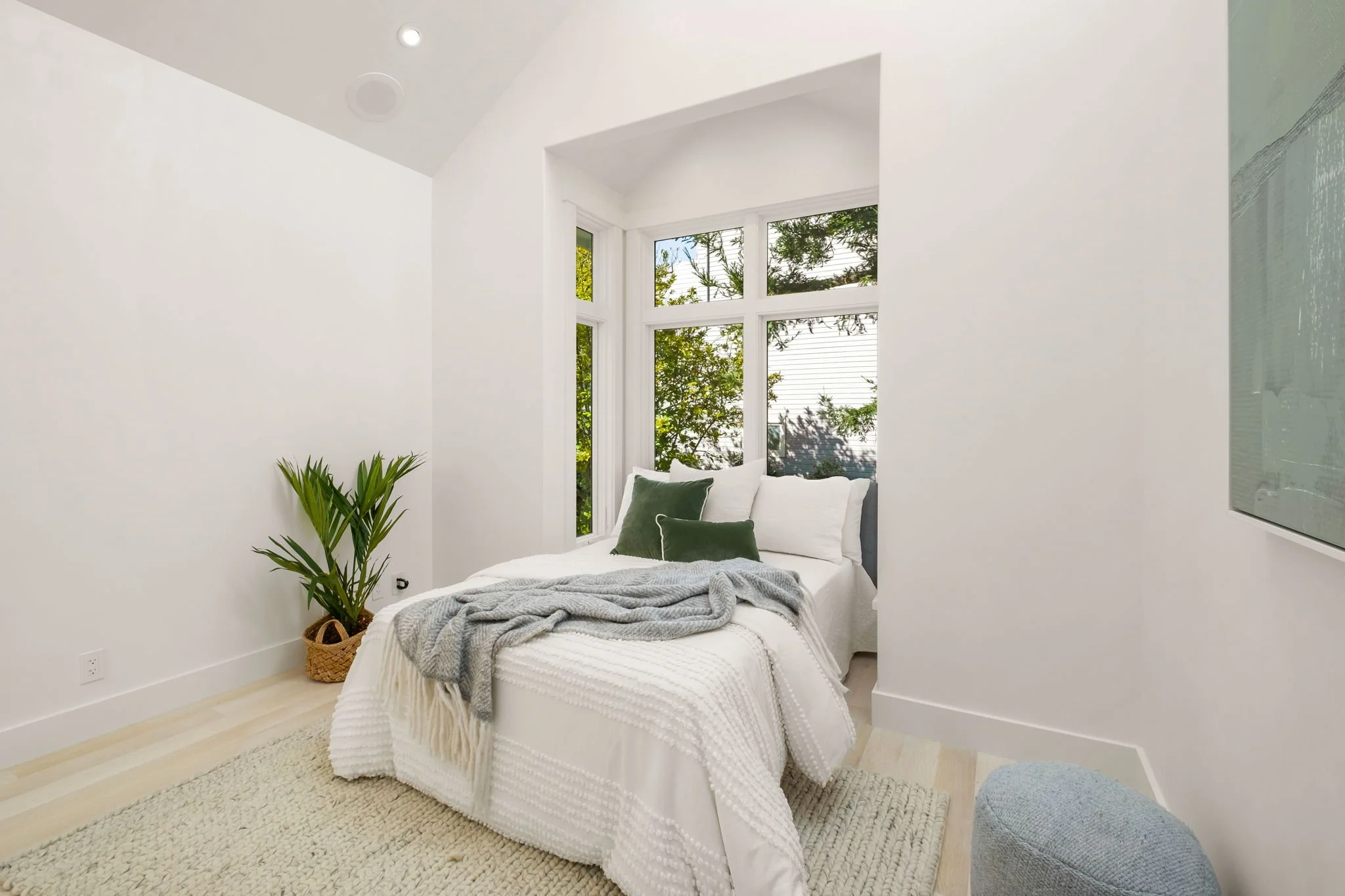 A small bedroom with white walls, a bed with white linens and gray blanket, green and white pillows, a window seat with view of trees outside, a potted plant in a woven basket, a beige rug, a light blue ottoman, and a poster on the wall.