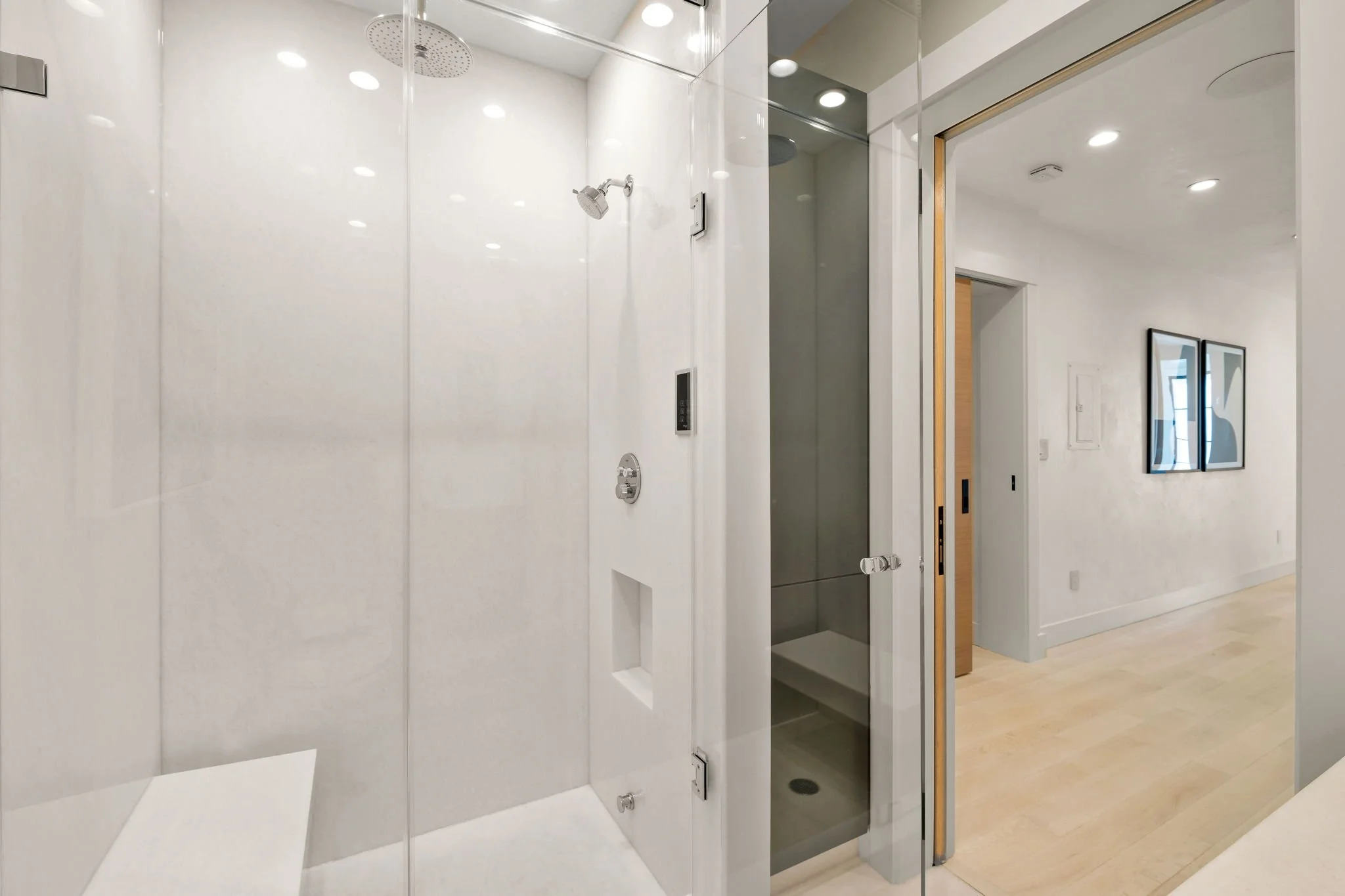 Modern bathroom with a glass-enclosed shower and a view of a hallway with light wood flooring and minimal artwork on the wall.