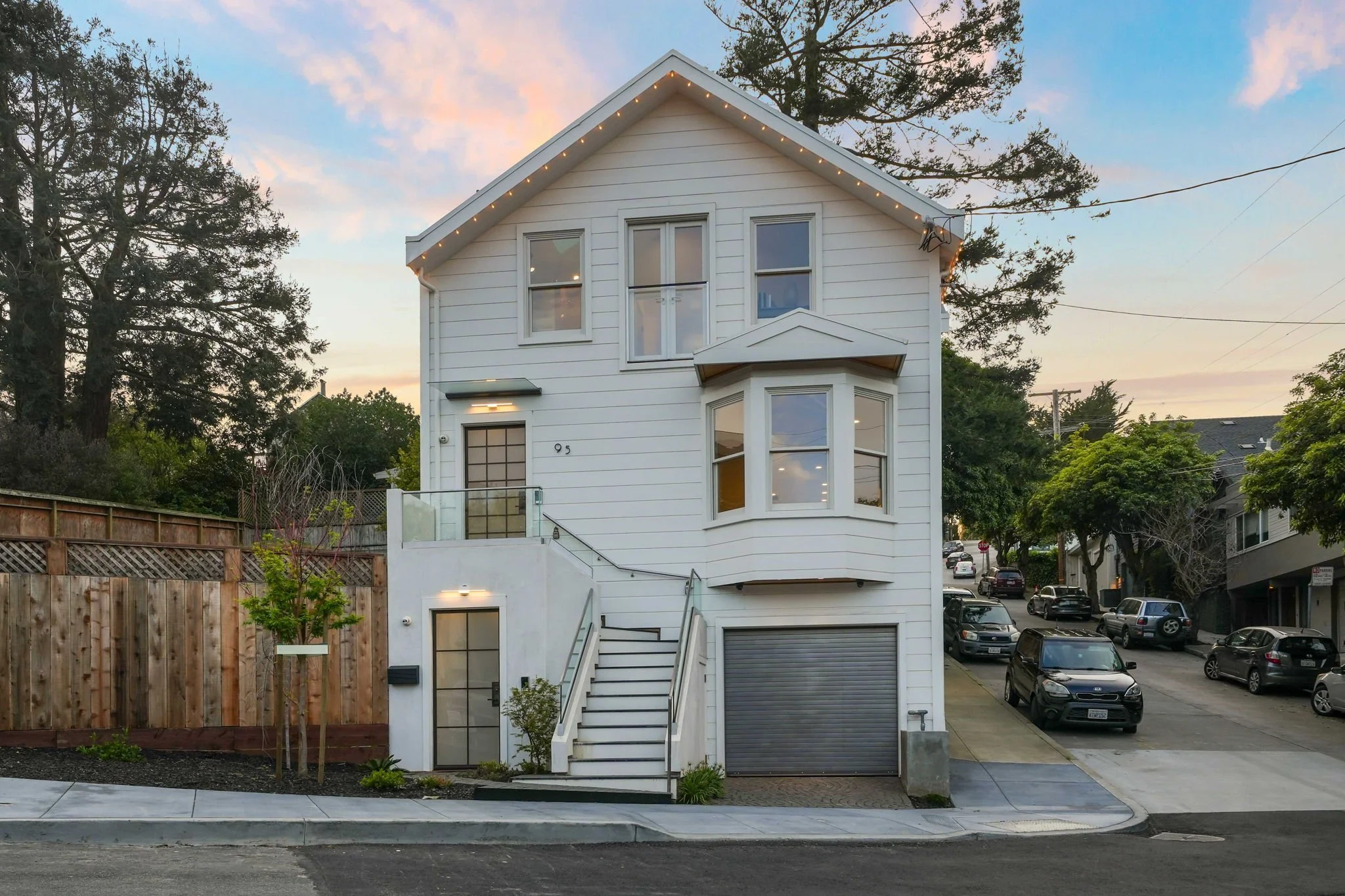 A modern three-story white house with a garage, exterior stairs, and multiple windows, situated on a city street with parked cars and trees.