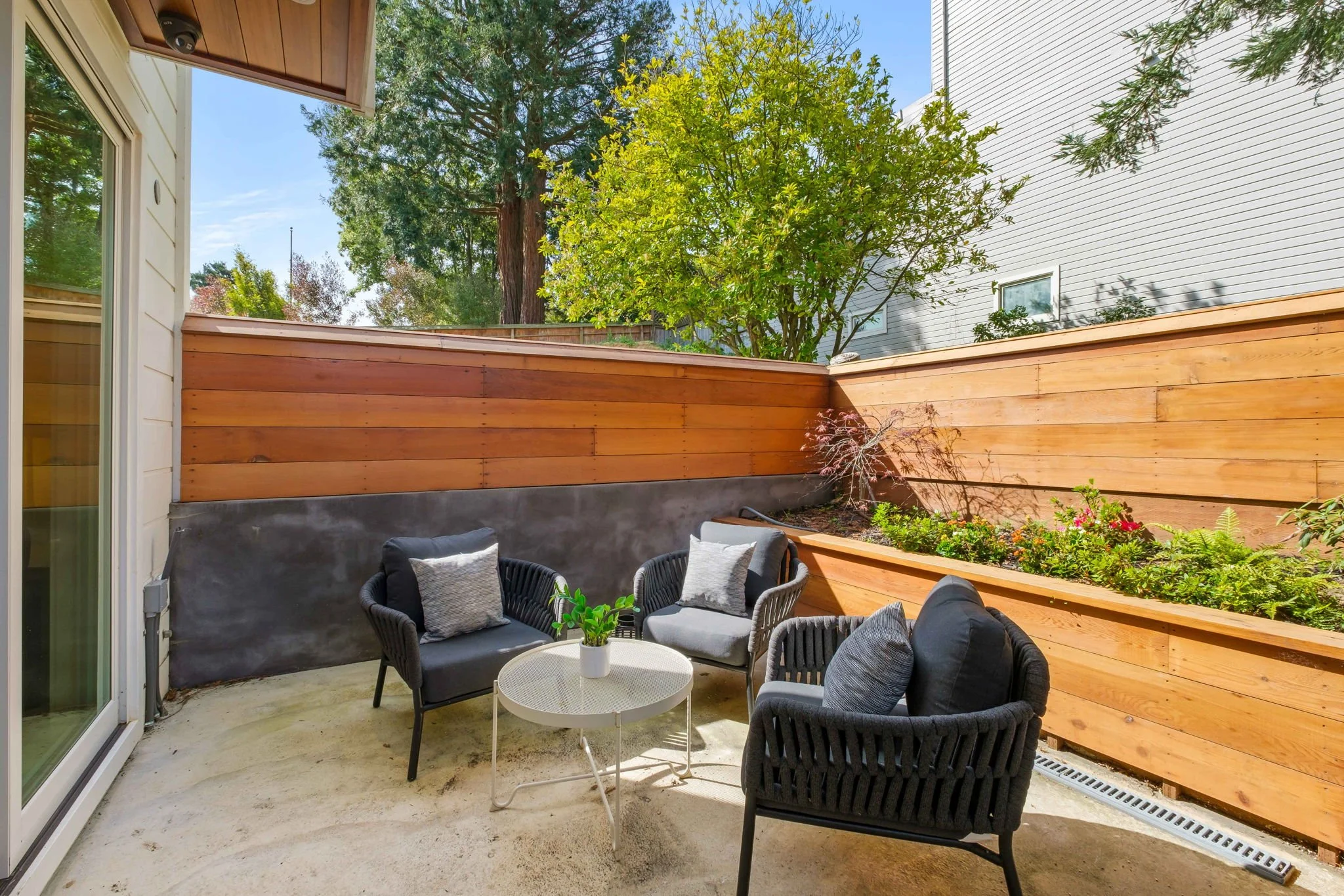 A cozy outdoor patio with three black woven chairs, each with a cushion, arranged around a white round table with a small plant in a white pot. The patio is enclosed by a wooden privacy fence with a planter bed containing plants and flowers, and a la