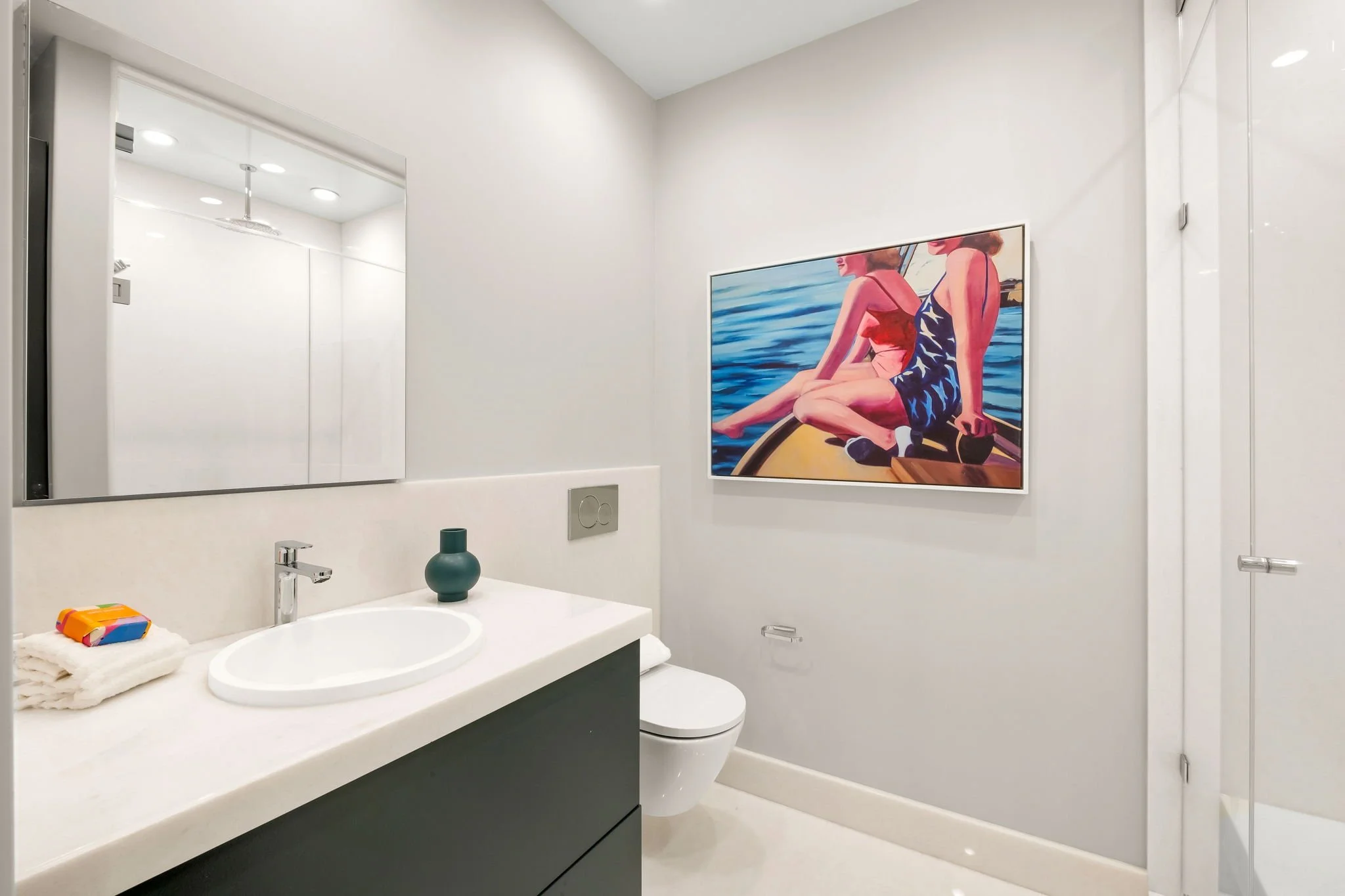 Modern white bathroom with a large mirror, a sink with a towel and soap on the counter, a green vase, a painting of women in swimwear by the water on the wall, and a toilet.