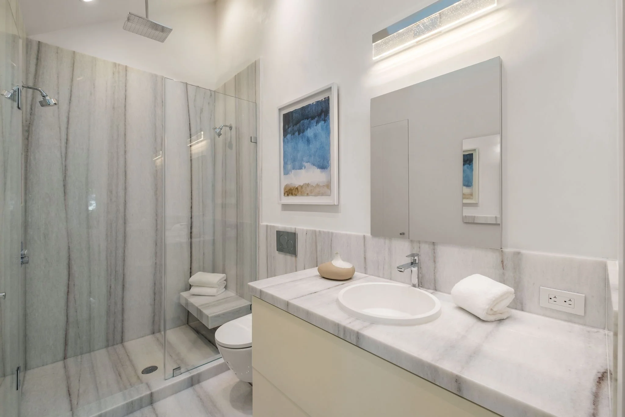 Modern bathroom with a marble countertop, a round sink, a large mirror, and a glass-enclosed shower with marble tiles. There is wall art featuring abstract blue, white, and brown colors, and bathroom accessories including rolled towels and a decorati