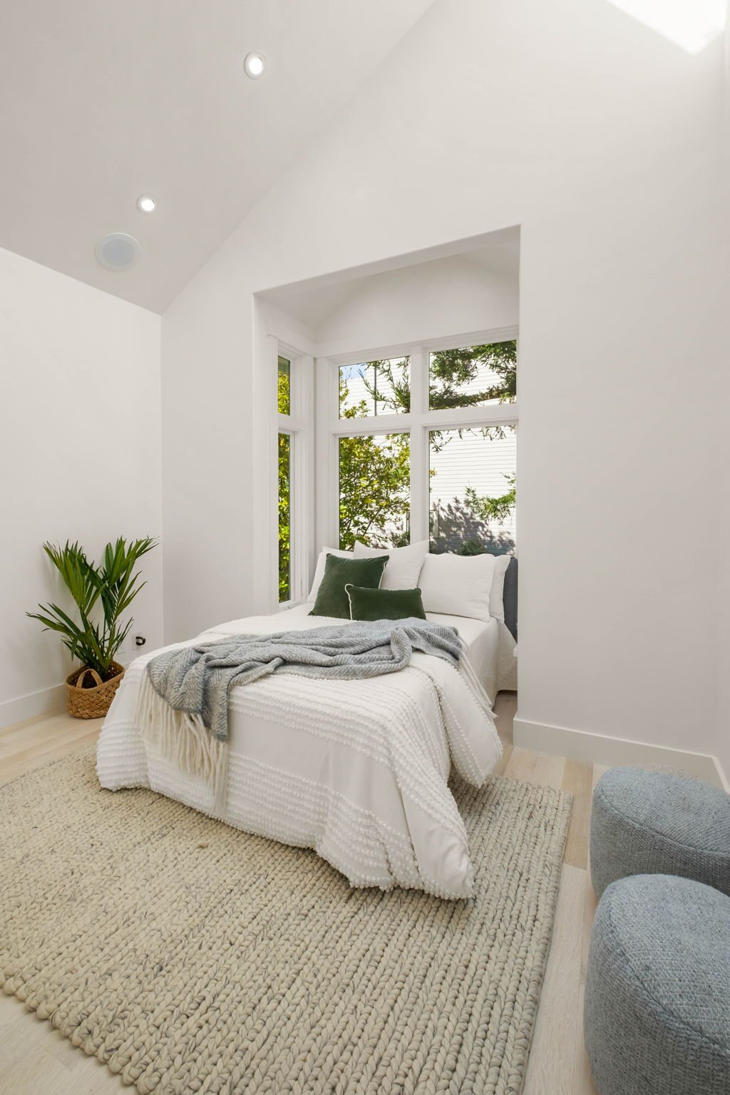 Bright bedroom with white walls and ceiling, featuring large windows with greenery outside, a white bed with green and white pillows, a gray blanket, a woven rug, a potted plant in the corner, and two gray poufs.