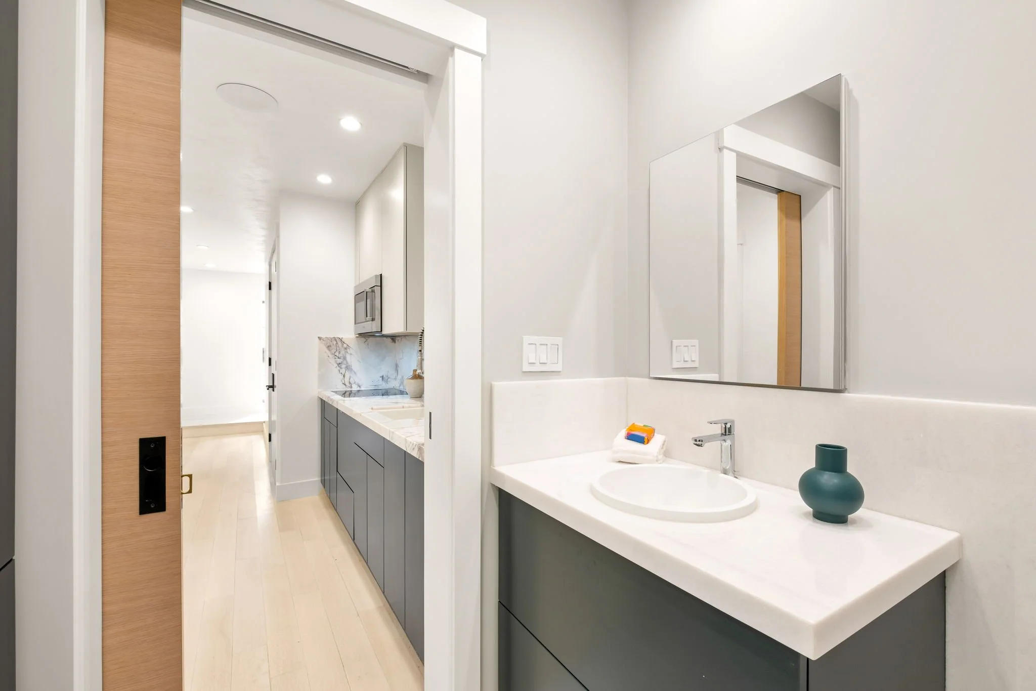 Modern bathroom with a white sink, green vanity, and a mirror, adjacent to a kitchen with gray and white cabinets and marble countertops.