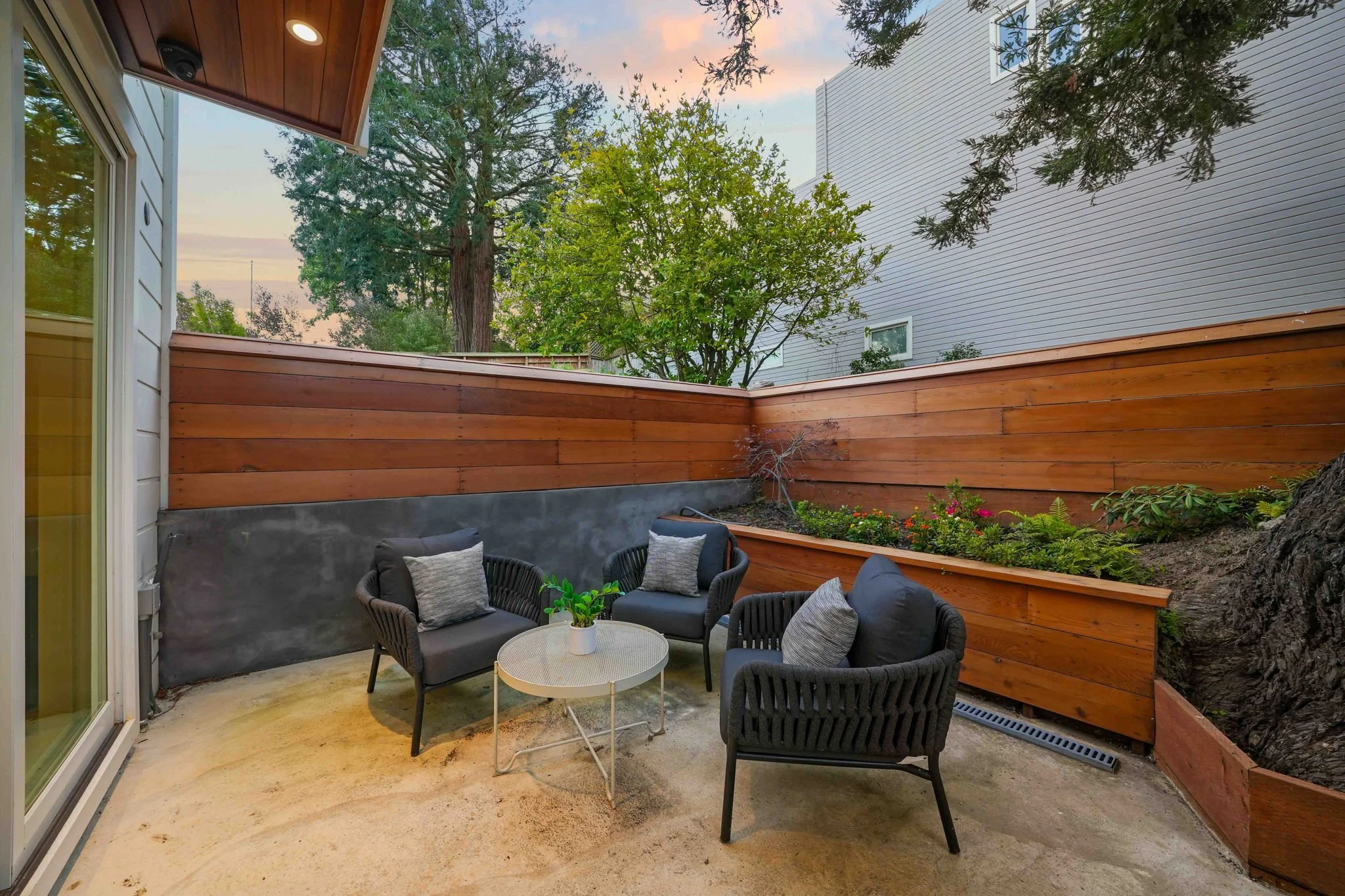 Small outdoor patio with three black chairs, a round table with a plant, wooden privacy fence, lush green trees, and plants in the background at dusk.