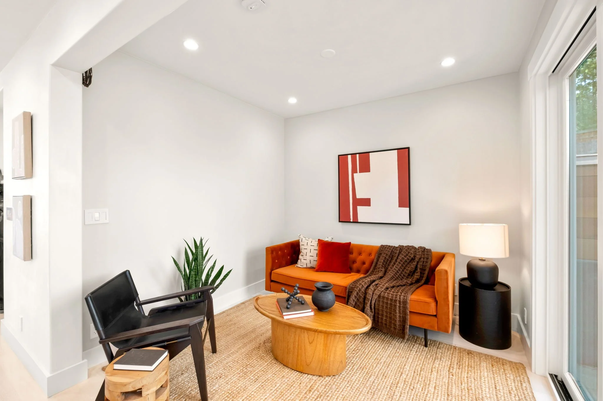 Living room with an orange sofa, black chair, round wooden coffee table, off-white walls, a red abstract painting, a white table lamp, a green plant, and a large window.
