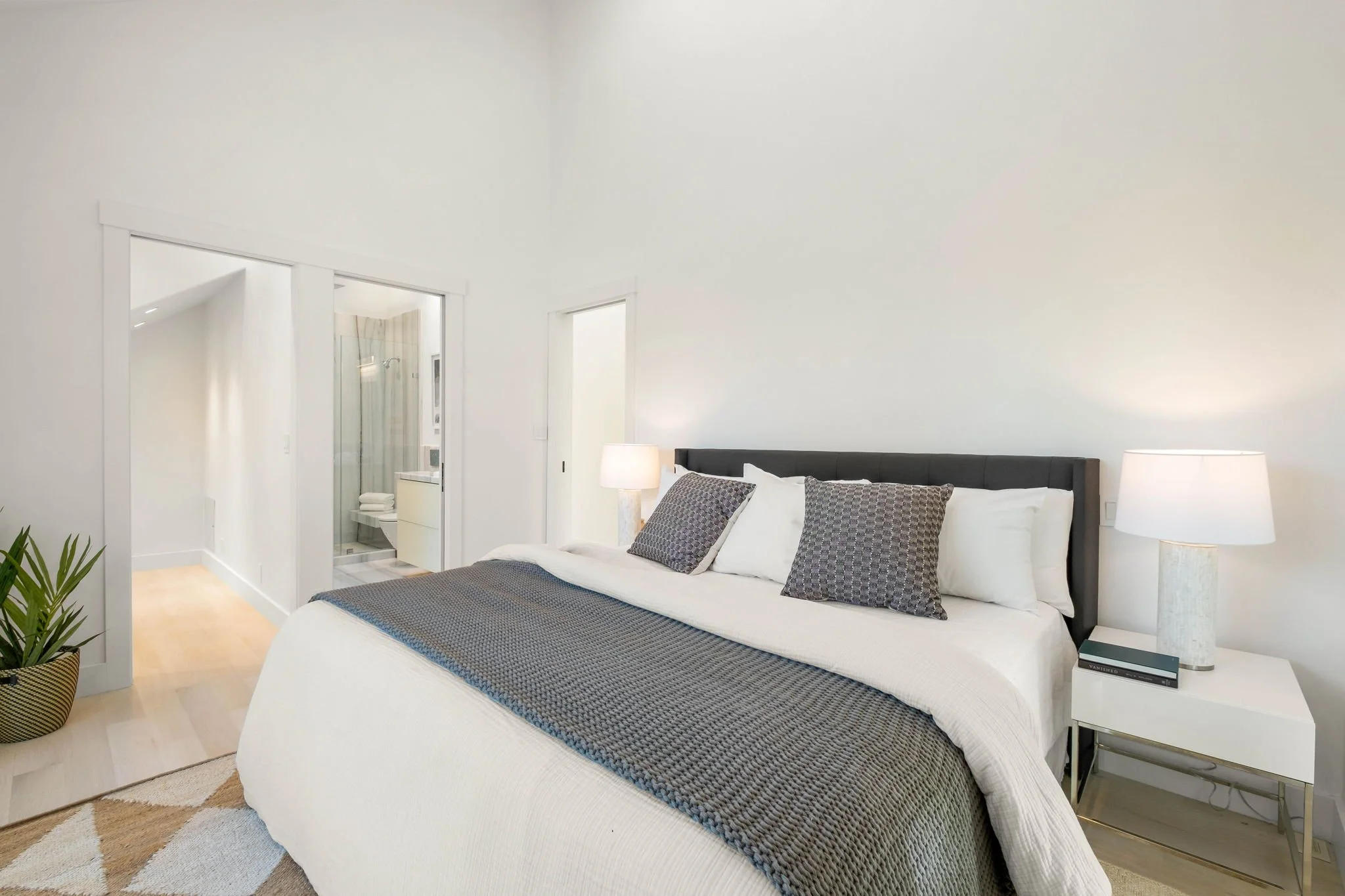 Modern bedroom with white walls, a large bed with dark gray headboard, white bedding, and patterned gray pillows. Nightstands with lamps on either side of the bed, and a closet with a sliding door leading to a bathroom.